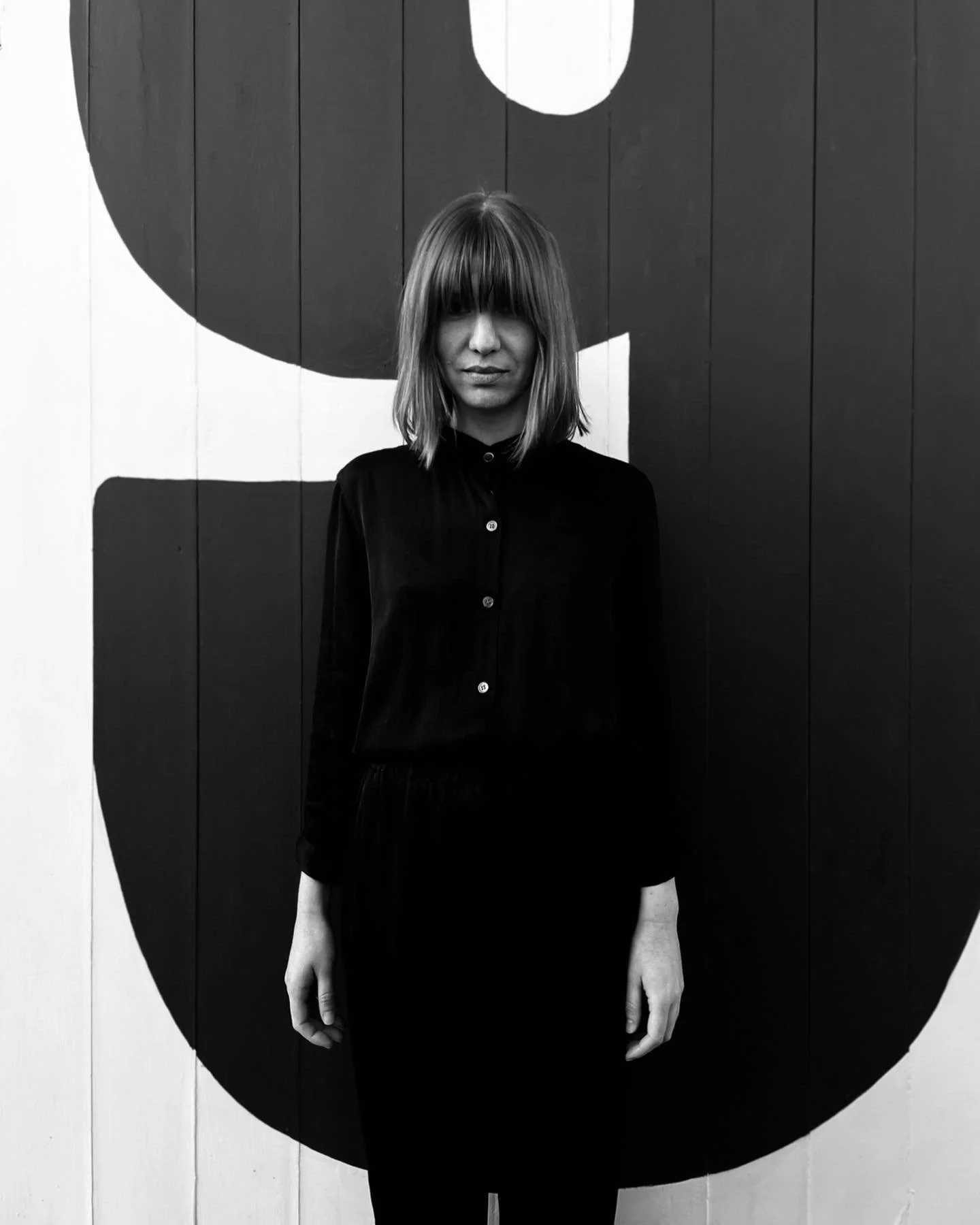 A woman with shoulder-length hair and bangs standing in front of a wall with a large, abstract animal face painted on it, in black and white.
