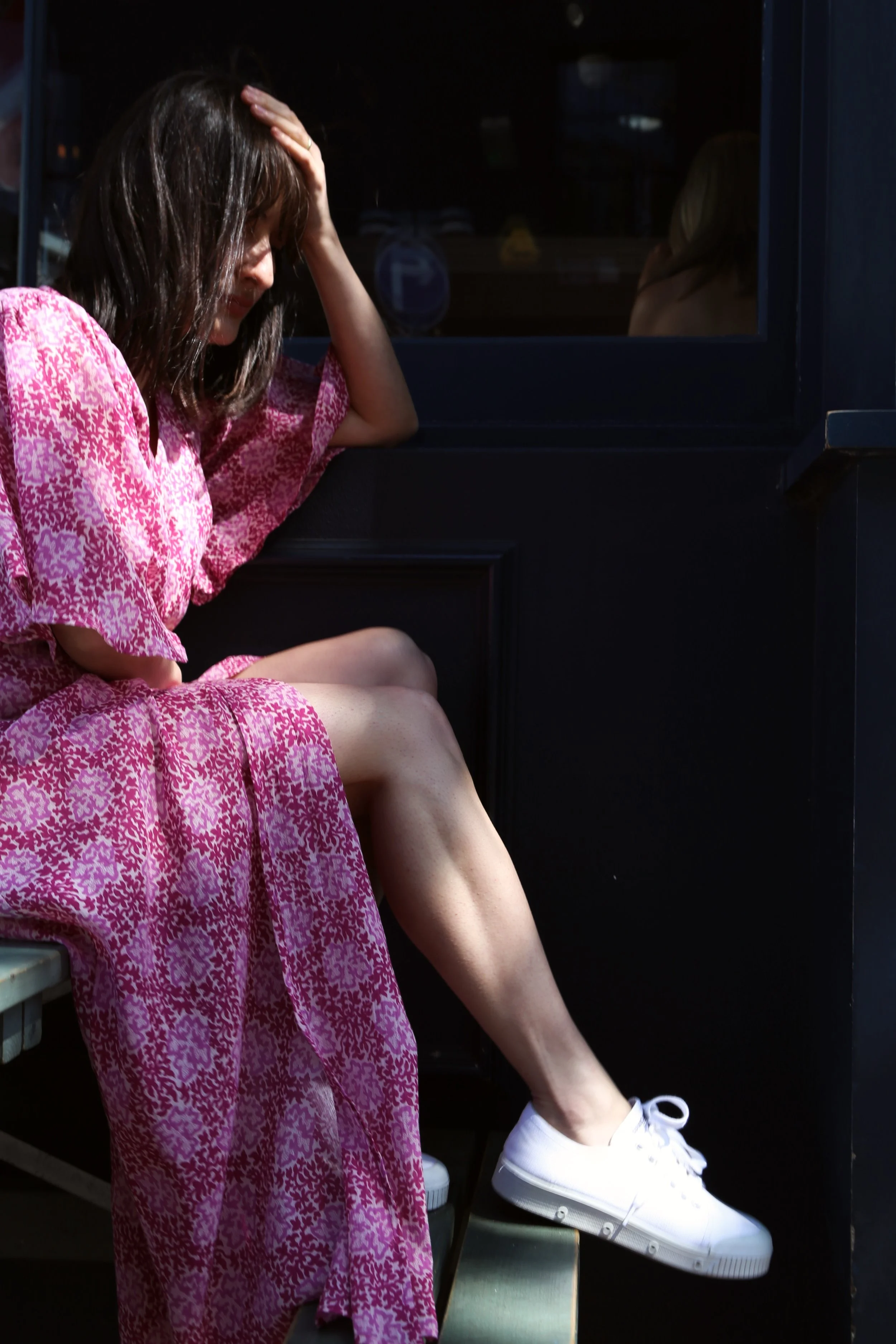 A woman wearing a pink patterned dress and white sneakers sitting with her hand on her forehead, appearing distressed or tired.