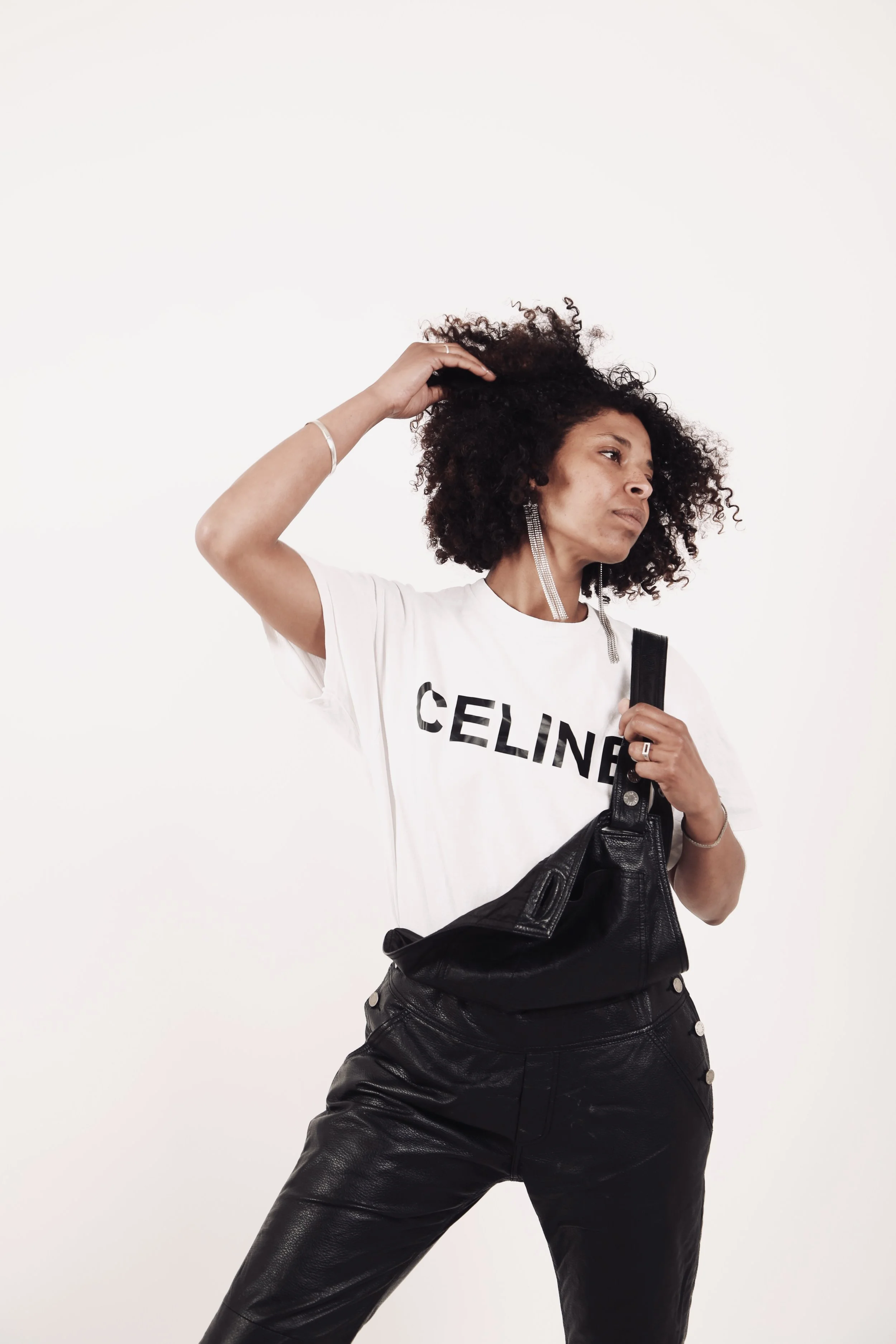 A woman with curly hair wearing a white t-shirt with 'CELINE' written on it, black leather pants, and large silver earrings, holding a black leather bag and touching her hair.