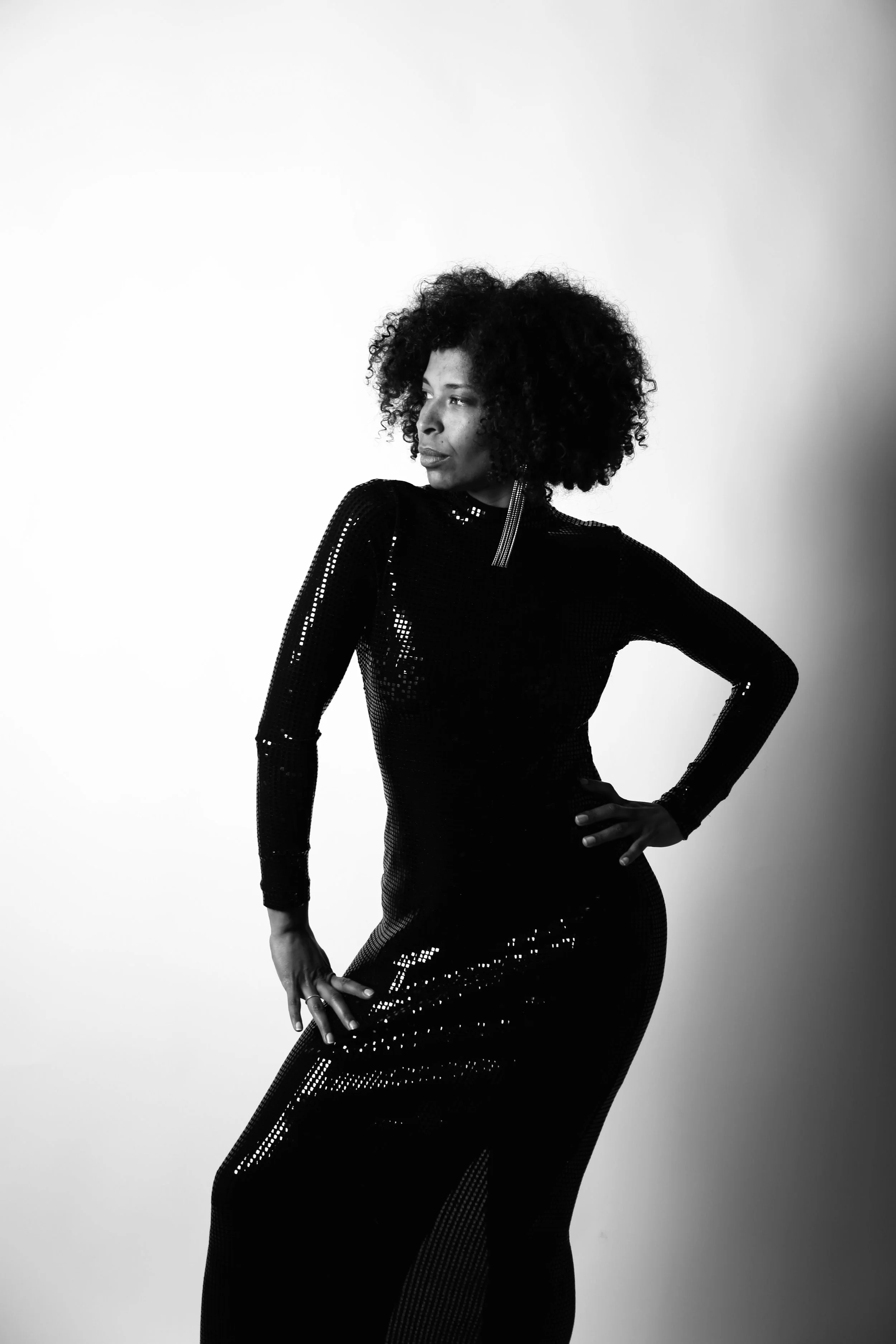 Black and white photo of a woman in a full-length dress with long sleeves and a high neck, posing with one hand on her hip against a plain background.