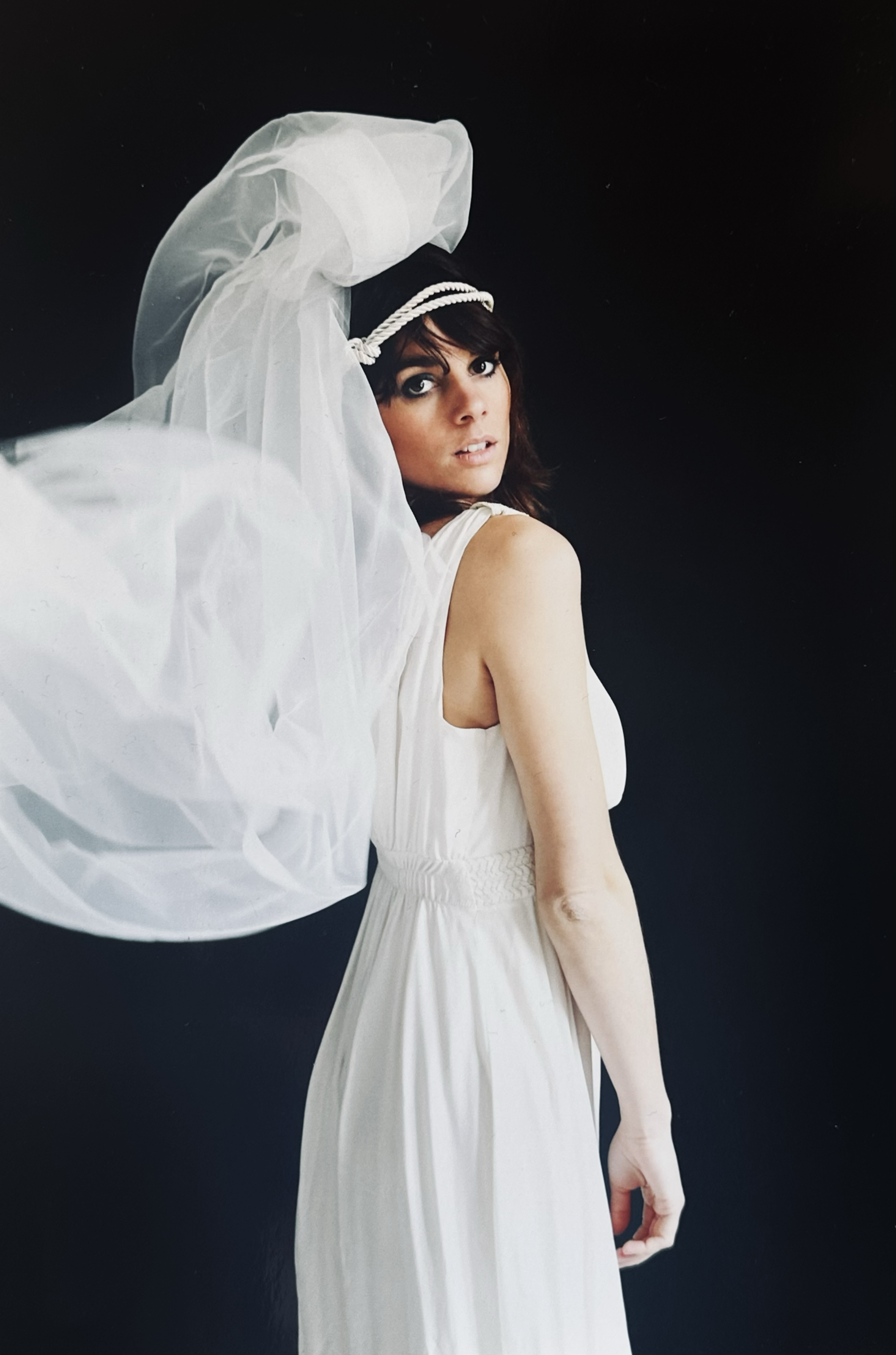 A woman in a white dress with a sheer veil and headband, standing against a dark background, looking over her shoulder.