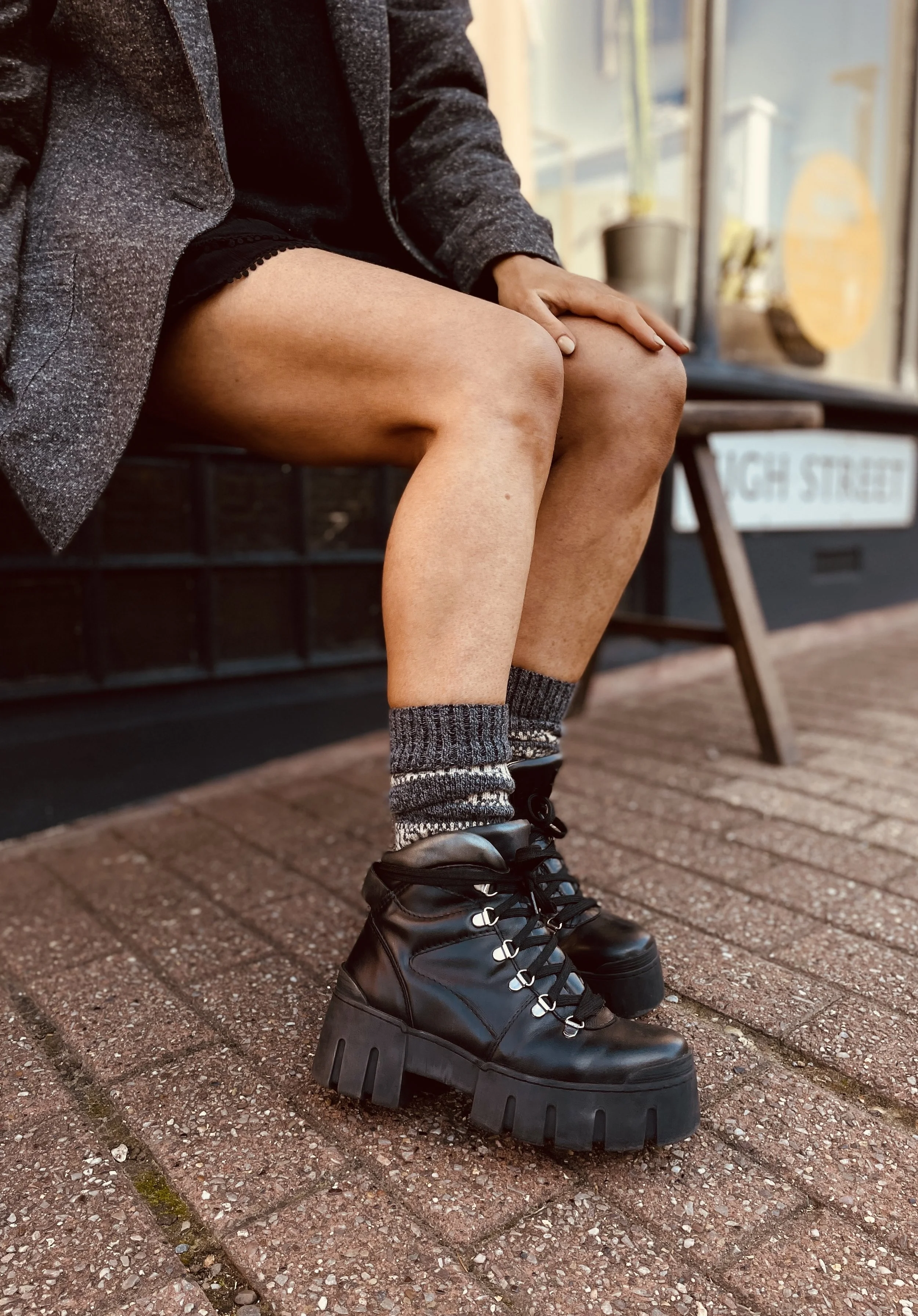 A person sitting on a bench wearing black chunky platform lace-up boots, patterned socks, and black shorts, with a gray blazer over their lap.