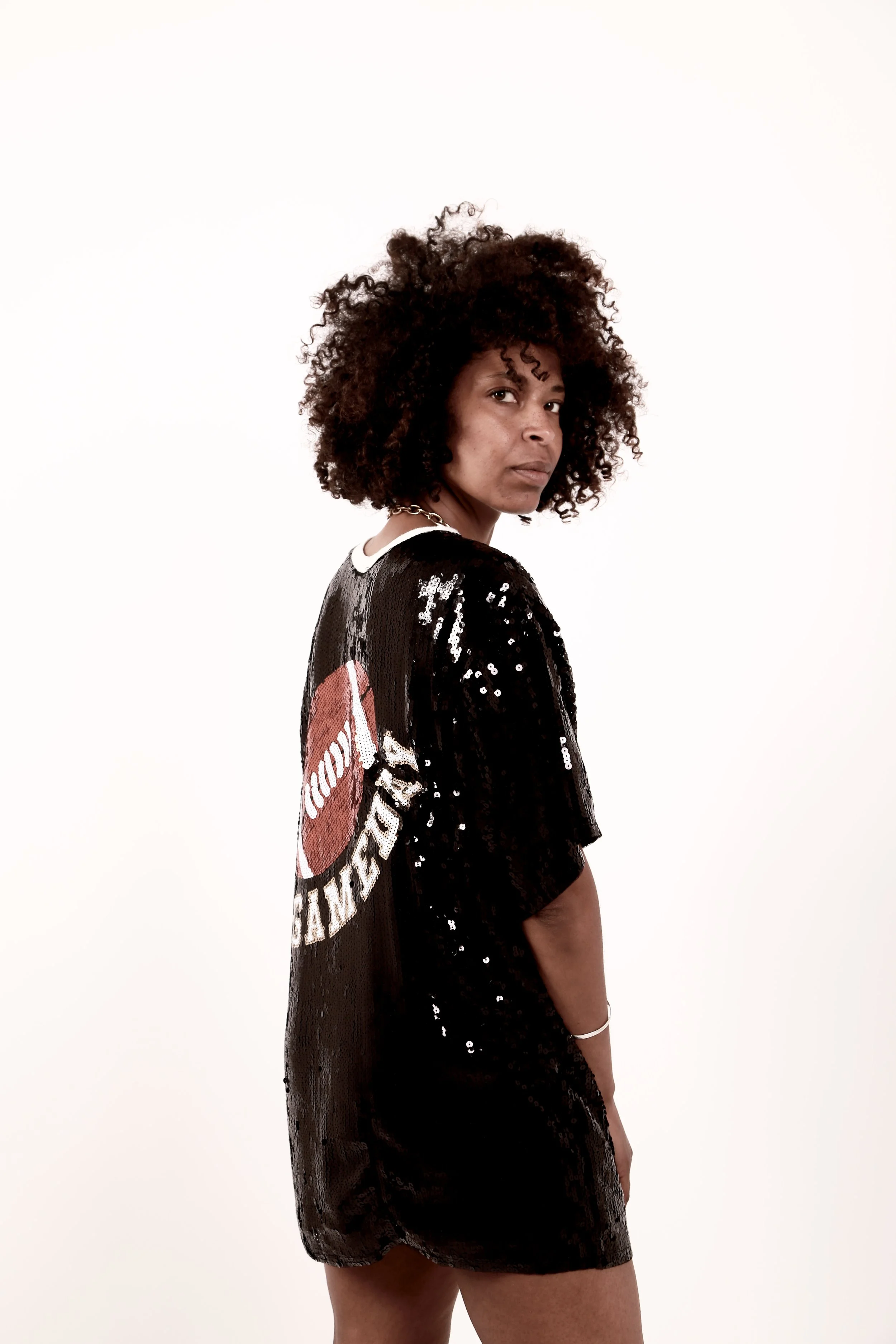A woman with curly hair wearing a black sequined dress with a patch on the back that reads 'SQUAD' and a red circle logo, standing against a plain white background.
