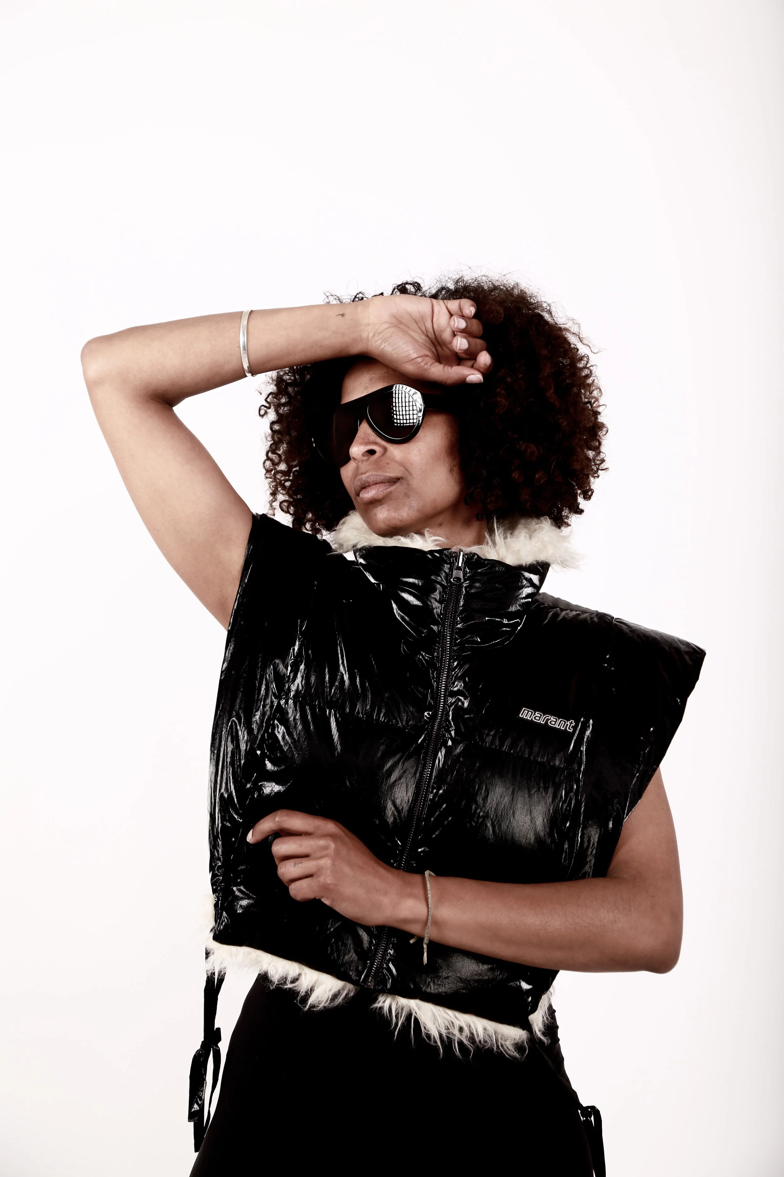 A woman with curly hair wearing sunglasses, a black shiny vest with a fur collar, and posing with her arm over her forehead.