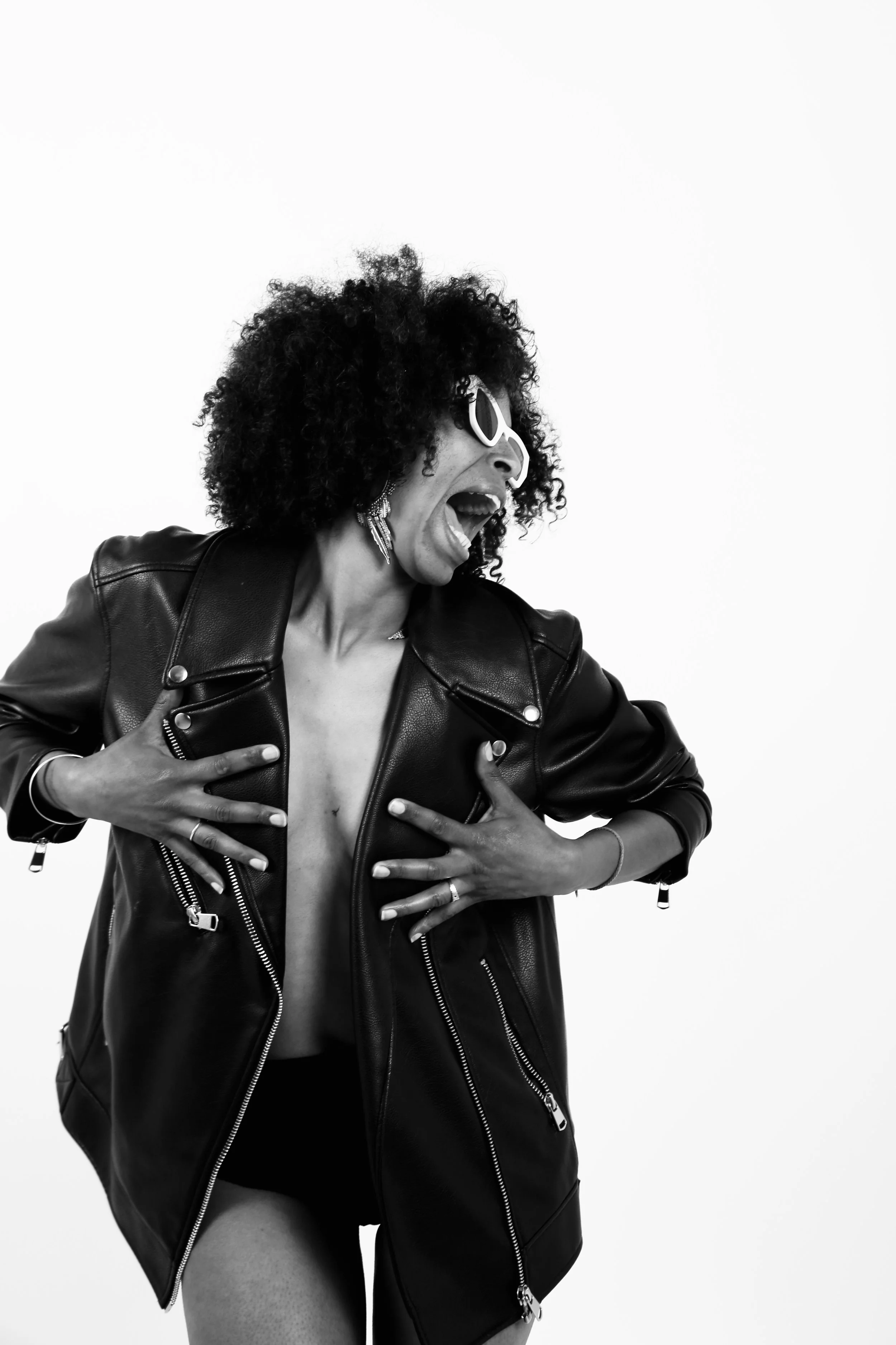 A woman with curly hair wearing a leather jacket, sunglasses, and jewelry, expressing excitement or joy with her mouth open, holding her jacket open to reveal her chest, against a plain background.