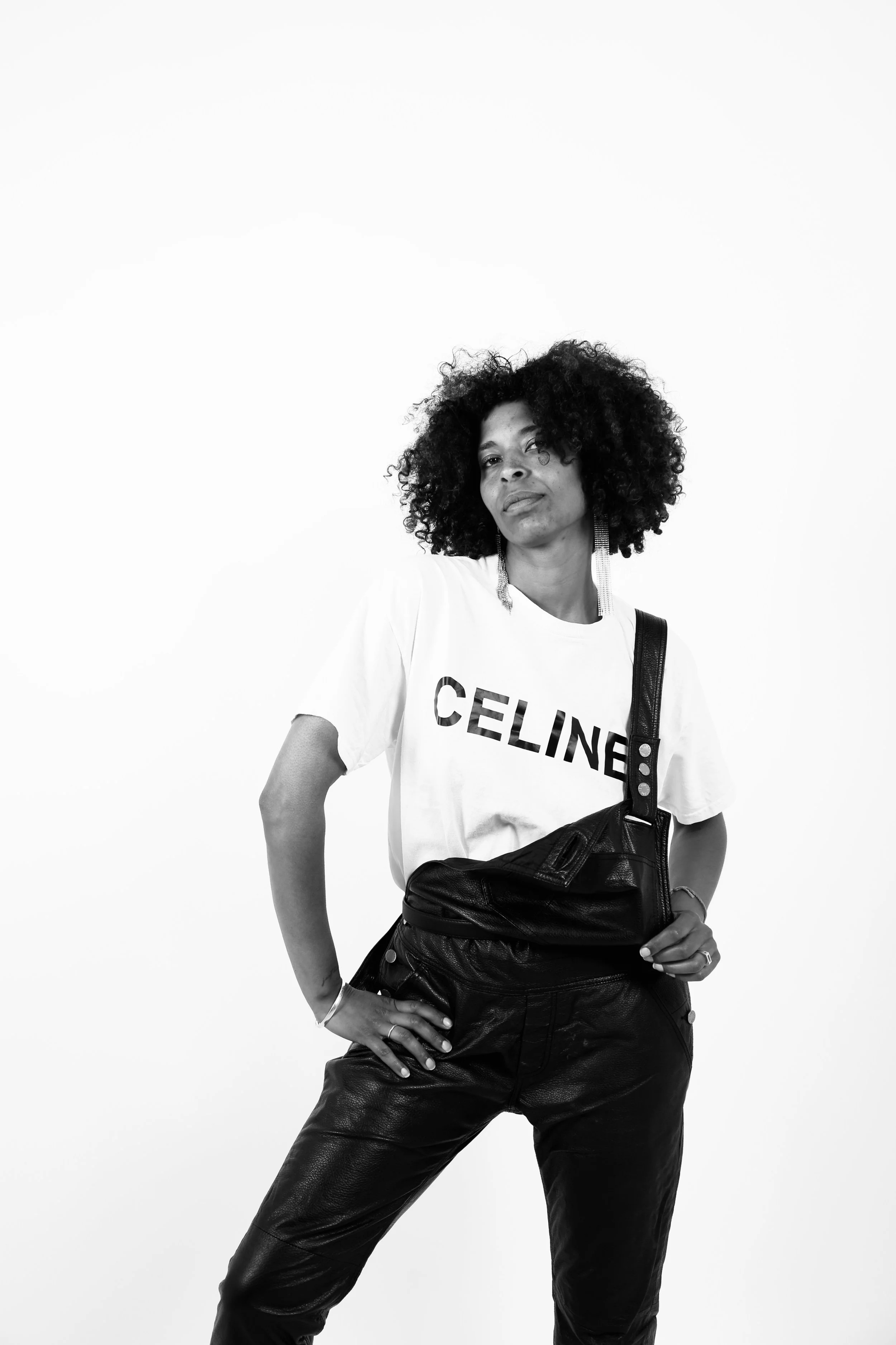 Black and white photo of a confident woman with curly hair, wearing a white T-shirt with 'CELINE' logo, leather pants, earrings, and carrying a black bag over her shoulder, standing against a plain background.