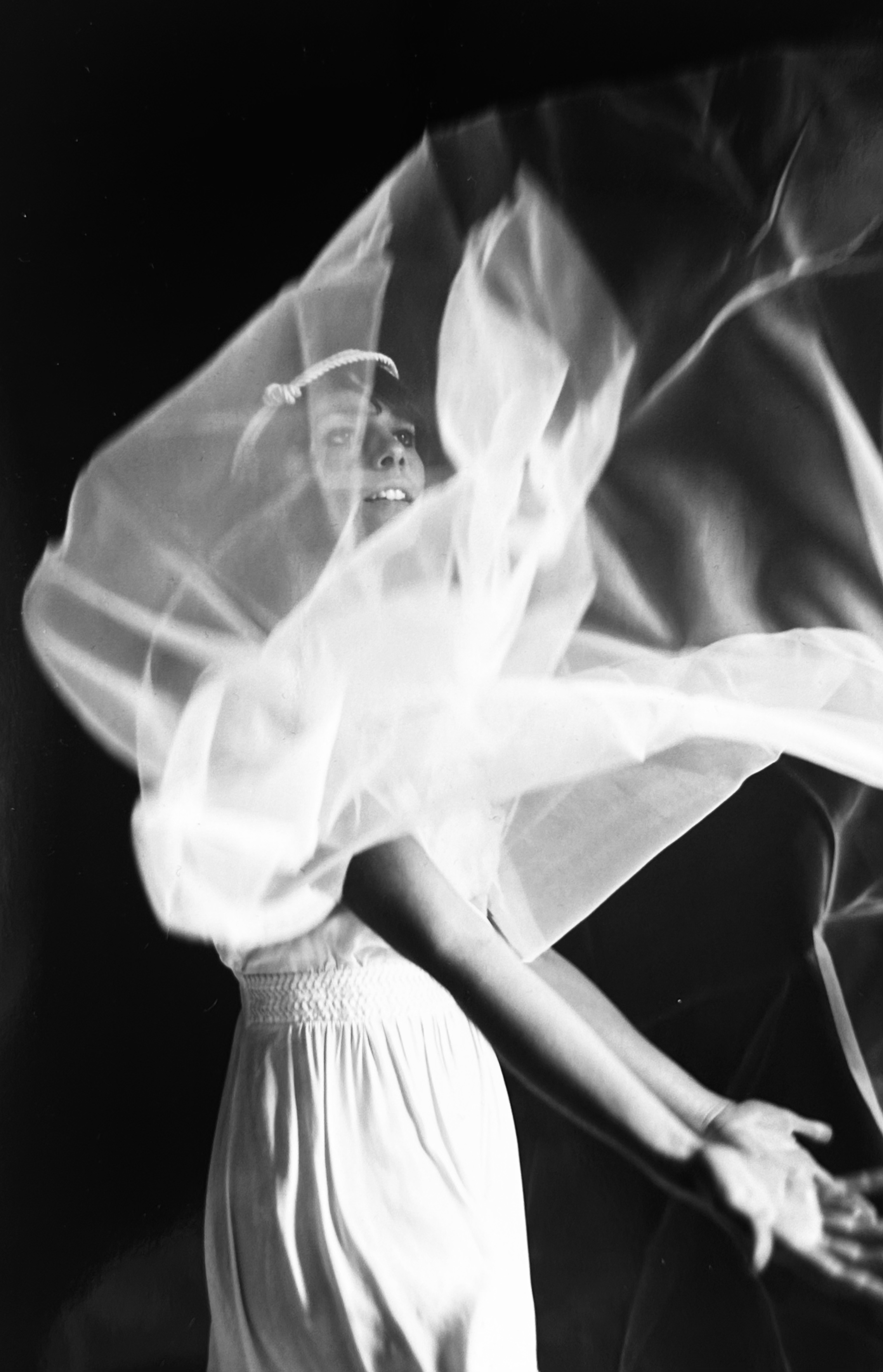 A woman smiling and looking to the side, wrapped in a flowing, translucent fabric, in a black and white photo.