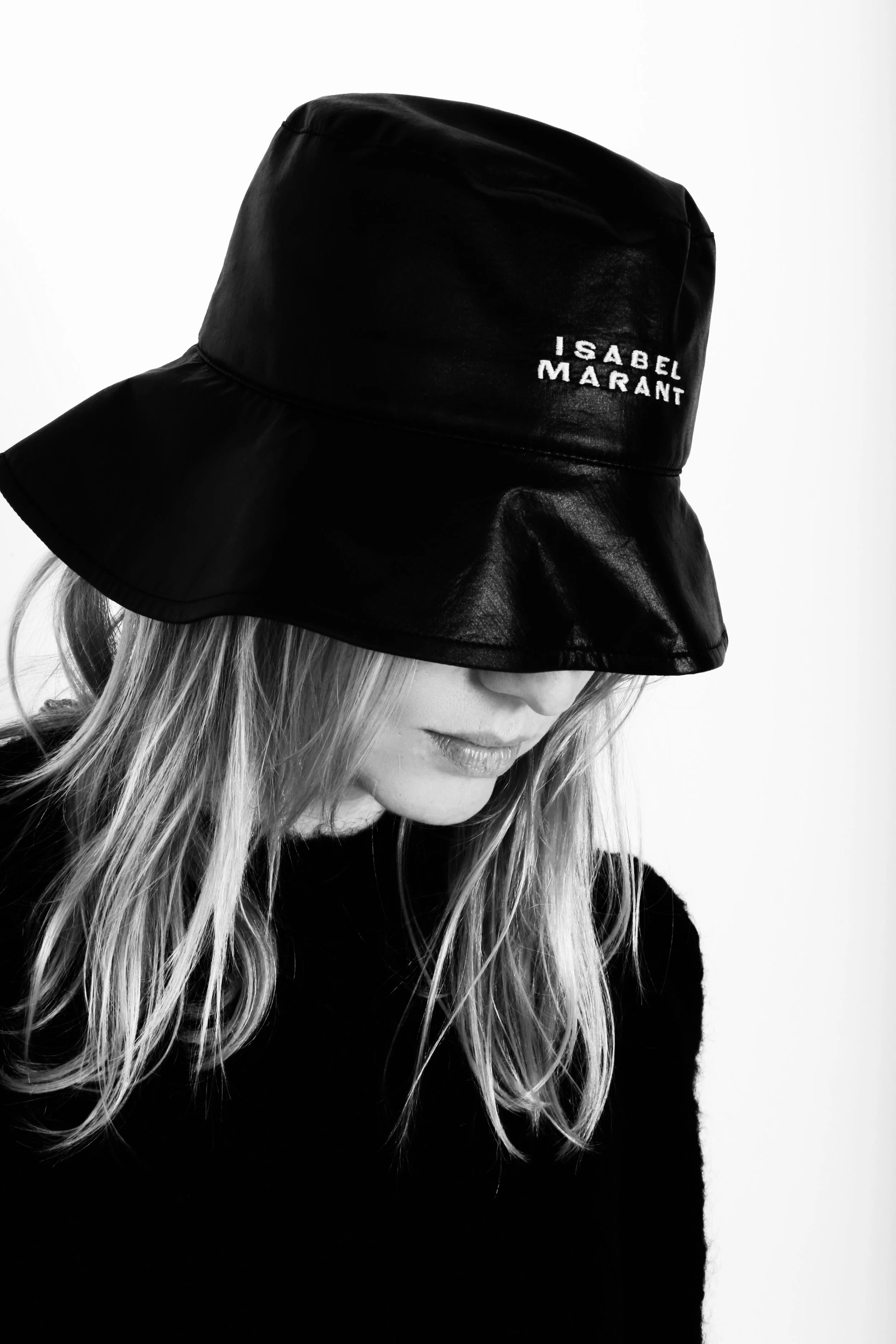 Black leather bucket hat with white text, worn slightly tilted downward by a woman with light hair, black top, in black and white photography.