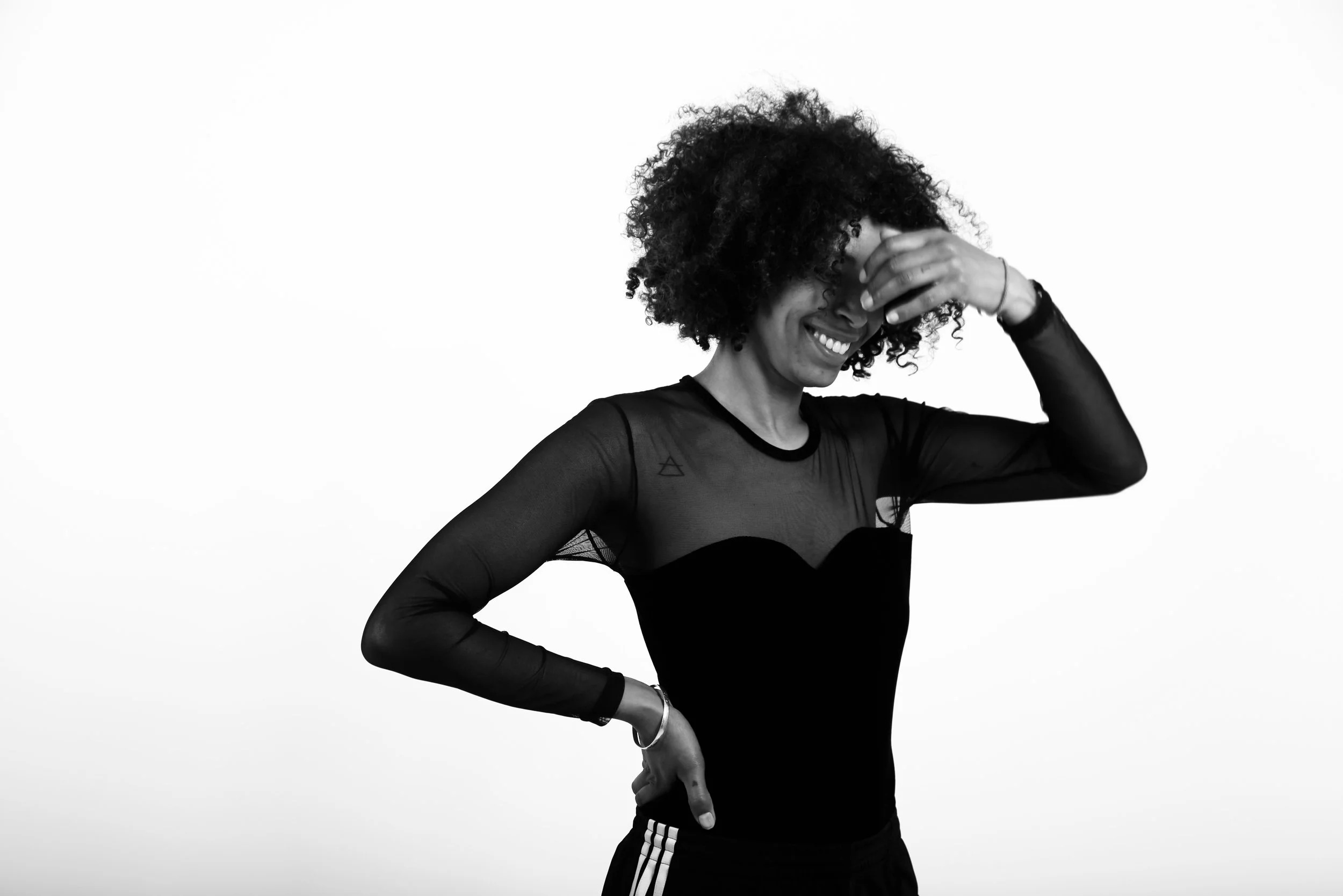 A woman with curly hair smiling and covering her eyes with her left hand, wearing a sheer long-sleeve top and black pants, standing against a plain white background.