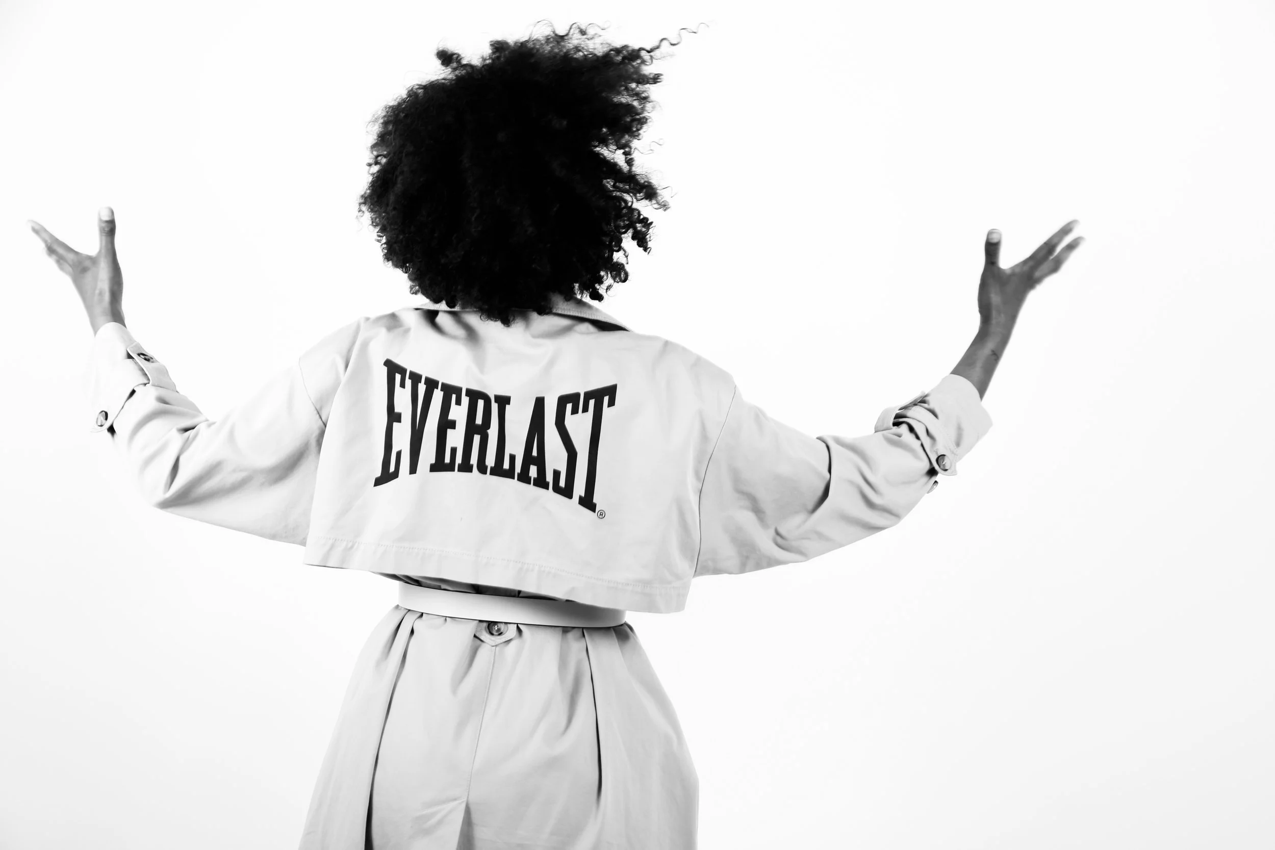 Back view of person with curly hair wearing a jacket with 'Everlast' logo, standing against a plain background with arms outstretched.