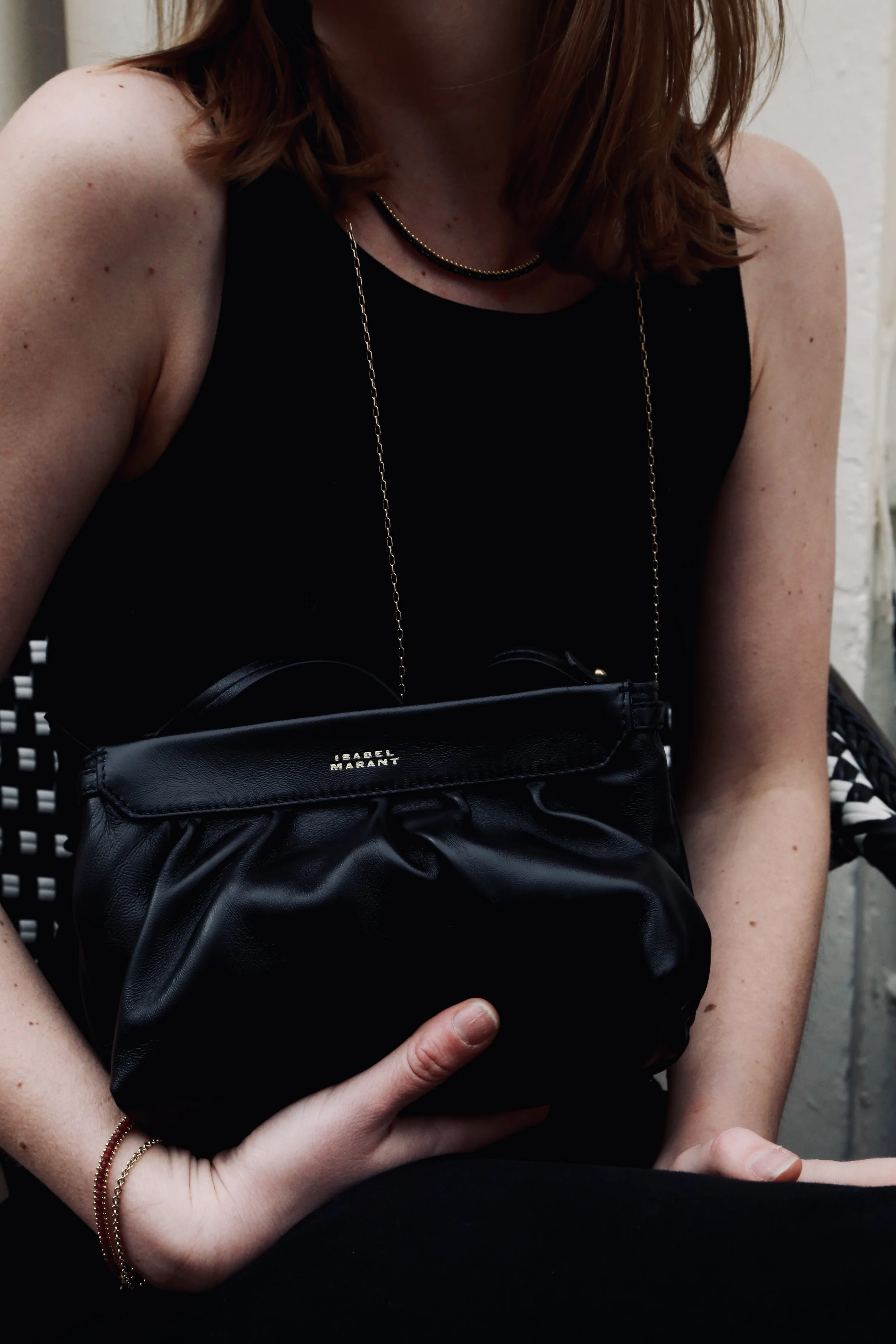 A woman wearing a black sleeveless top and gold necklaces holding a black handbag with a chain strap, with the brand name 'Isabel Marant' visible on the bag.