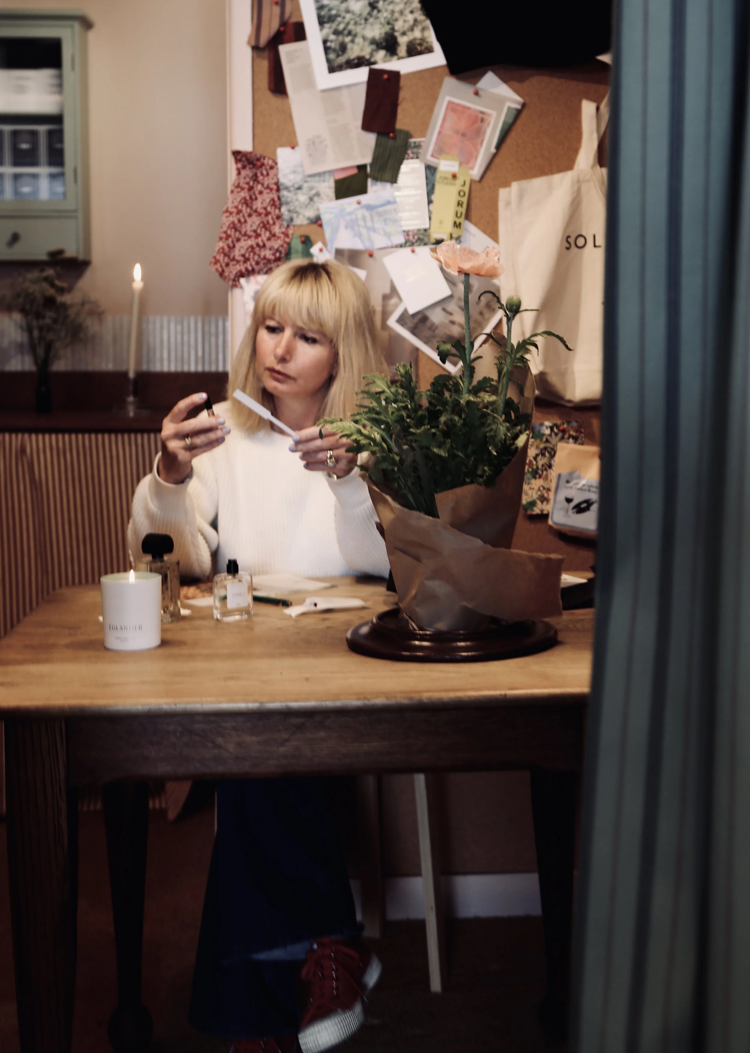 Woman sitting at a wooden table, applying perfume or makeup, with a potted plant, candle, and bottles on the table. The background features a bulletin board with photos and notes.