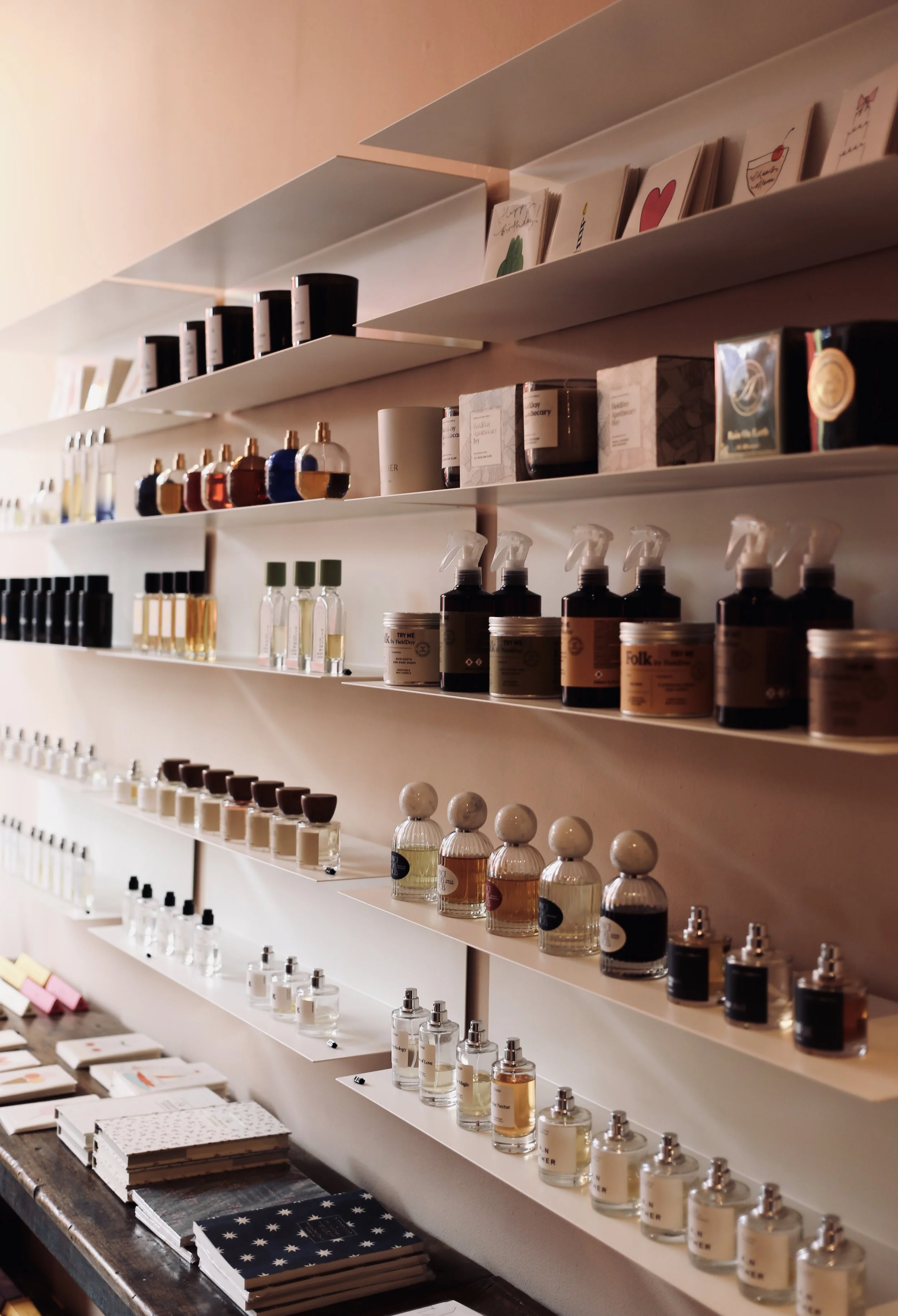 Shelves filled with various bottles and containers of fragrances, perfumes, and skincare products, with some notebooks or cards on a table below.