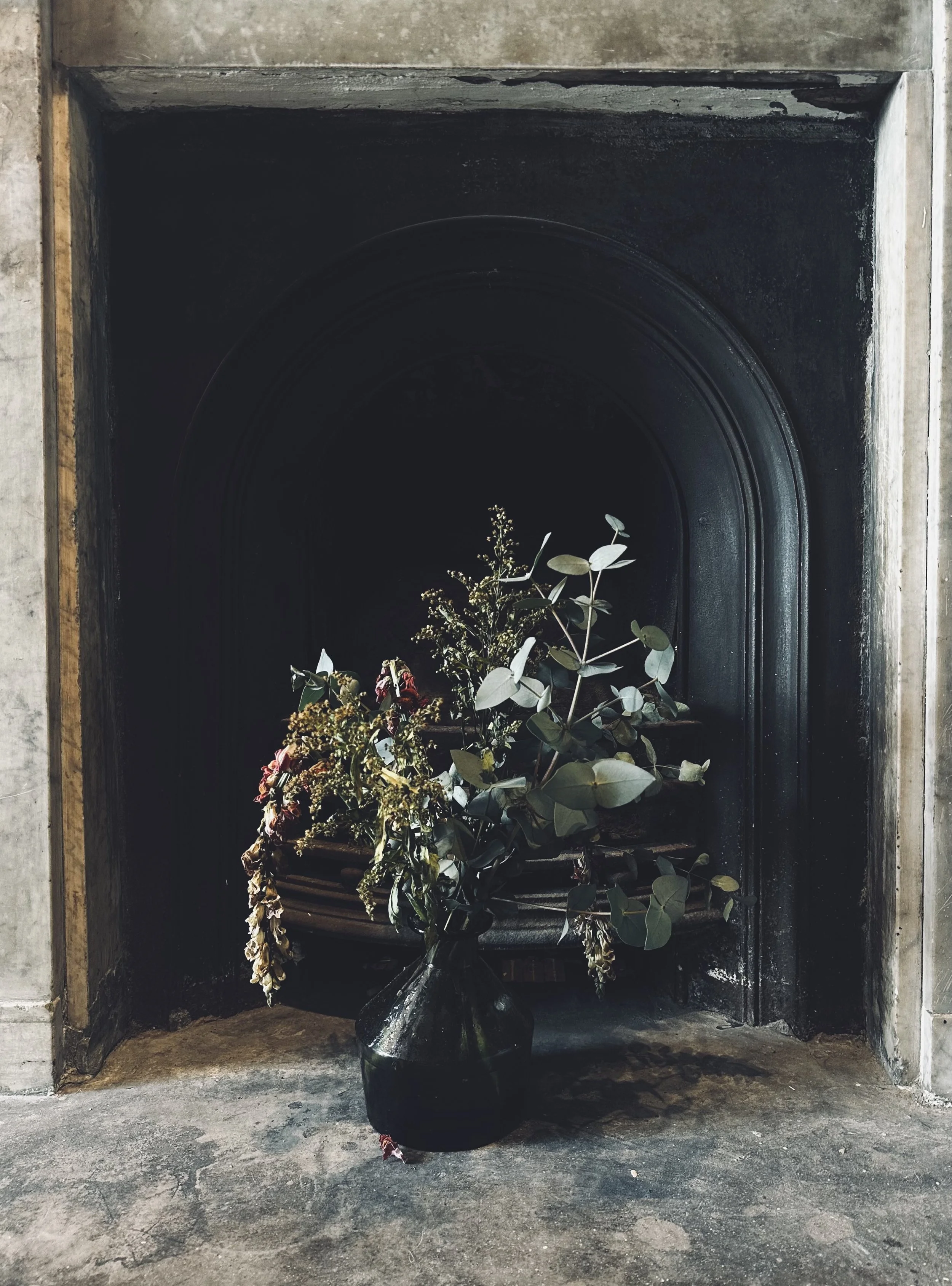 A black vase with green leaves and dried flowers, placed in front of a dark fireplace with a gray stone surround.