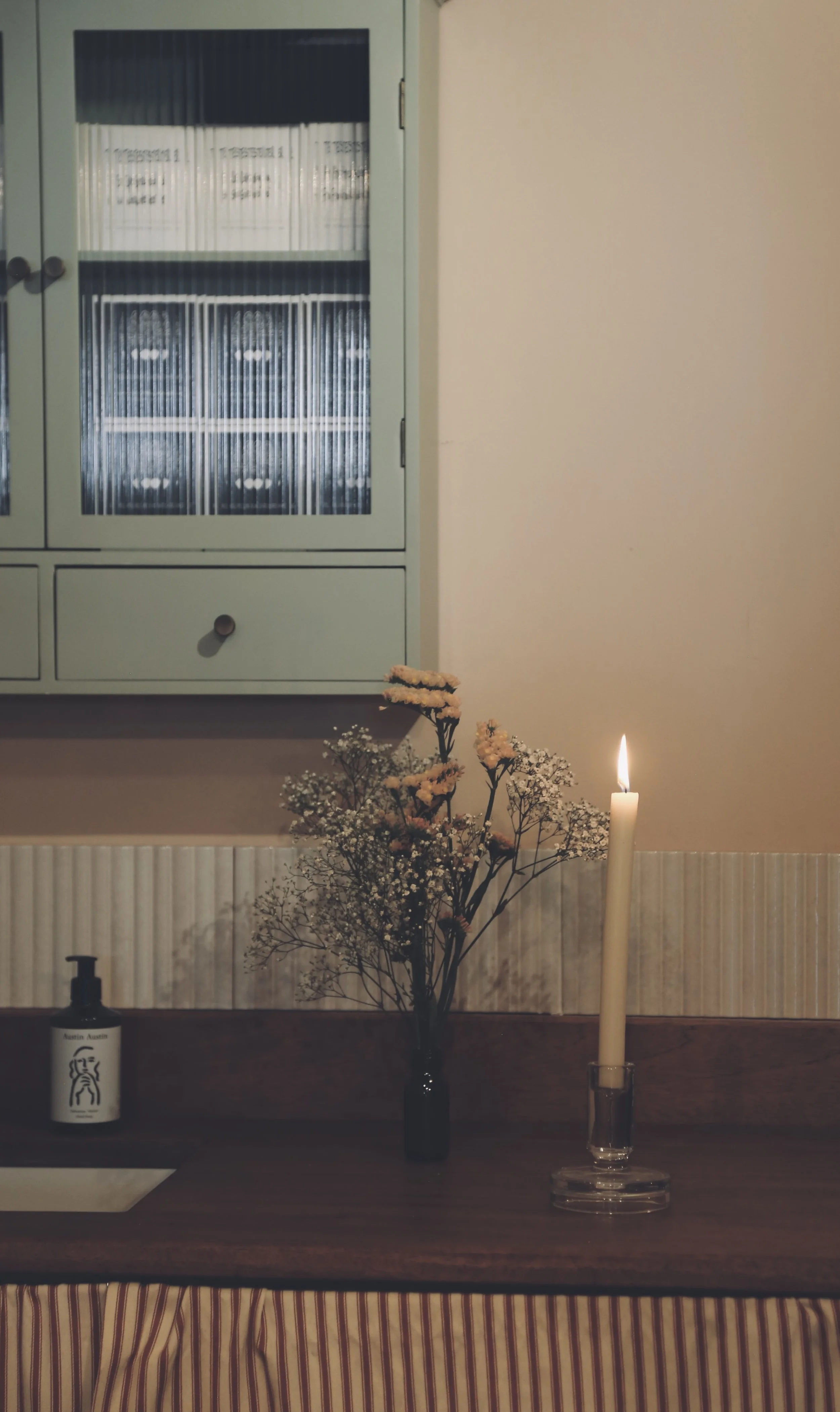 A small interior space with a dark wooden countertop, a black vase with white and peach flowers, a lit candle in a clear glass holder, a bottle of Austin Austin hand soap, a beige wall with a white radiator at the bottom, and a light green cabinet wi