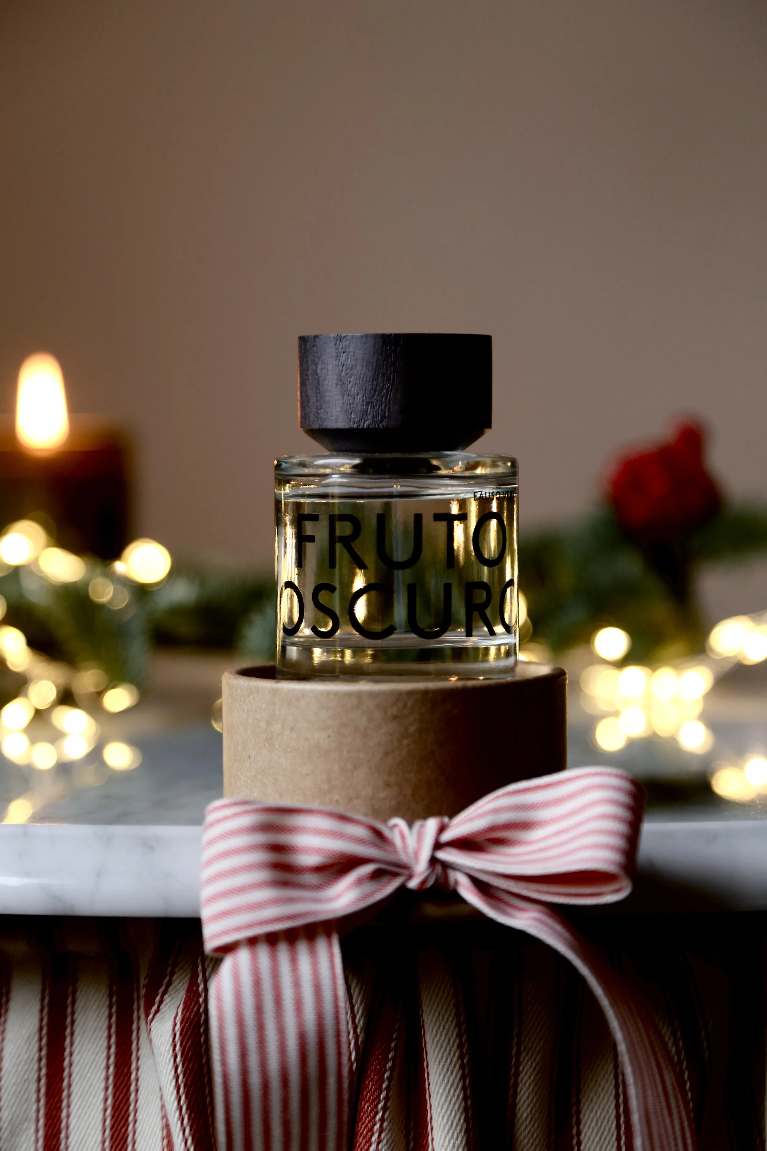 A small glass bottle labeled 'FRUTO OSCURO' is placed on a round cardboard box tied with a pink and white striped ribbon. The background features blurred holiday lights and a candle.
