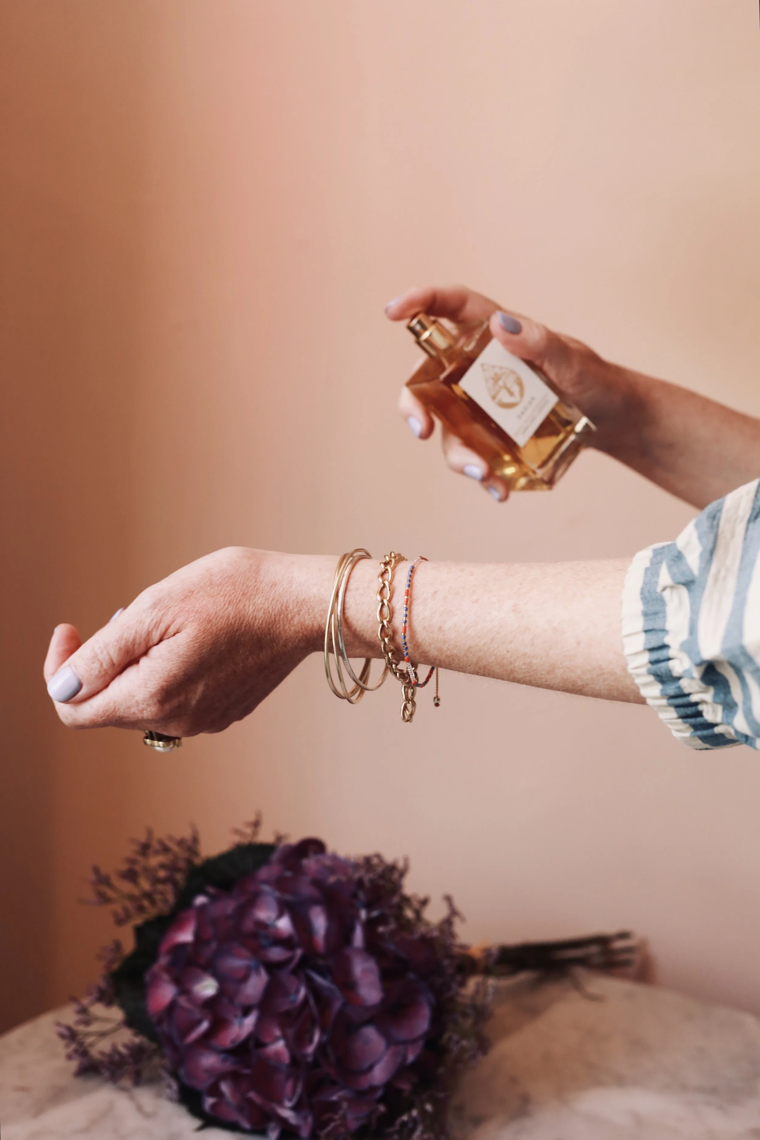 A person holding a perfume bottle with their right hand, wearing several gold bracelets, with a purple flower bouquet in the background on a surface.