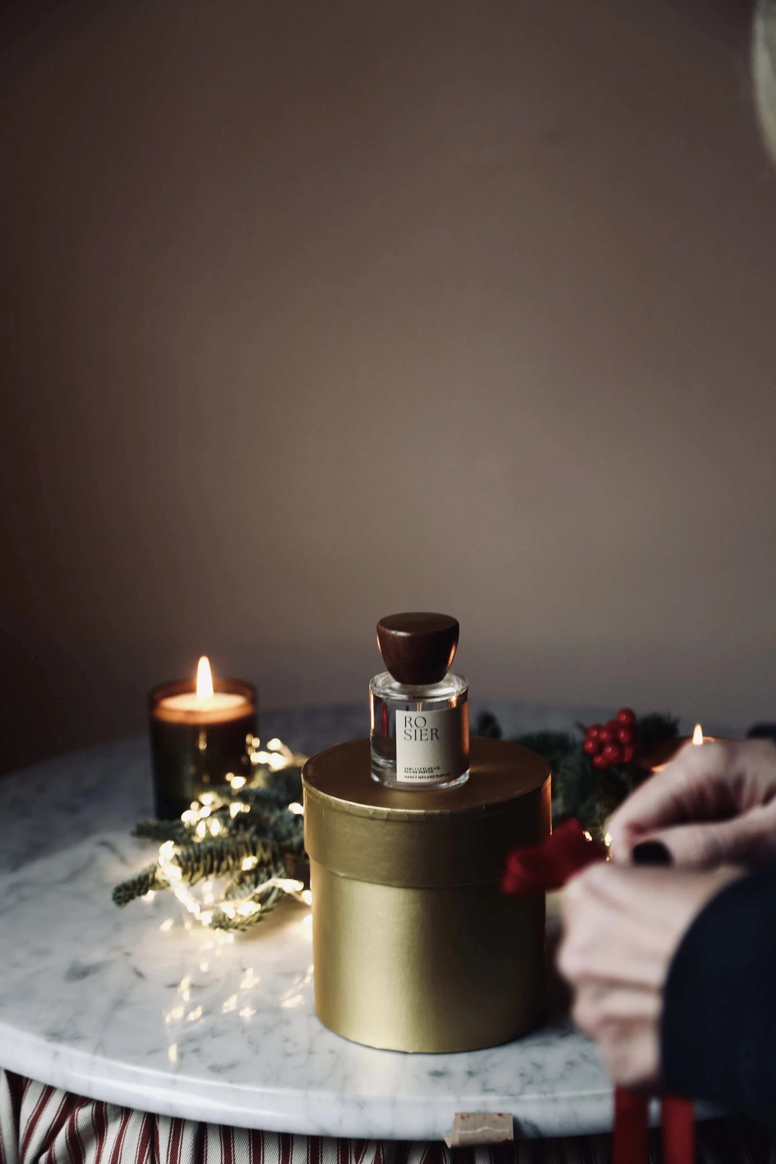 A small candle and a jar of perfume placed on a gold box on a marble table, with holiday decorations and a person's hand holding a red object nearby.