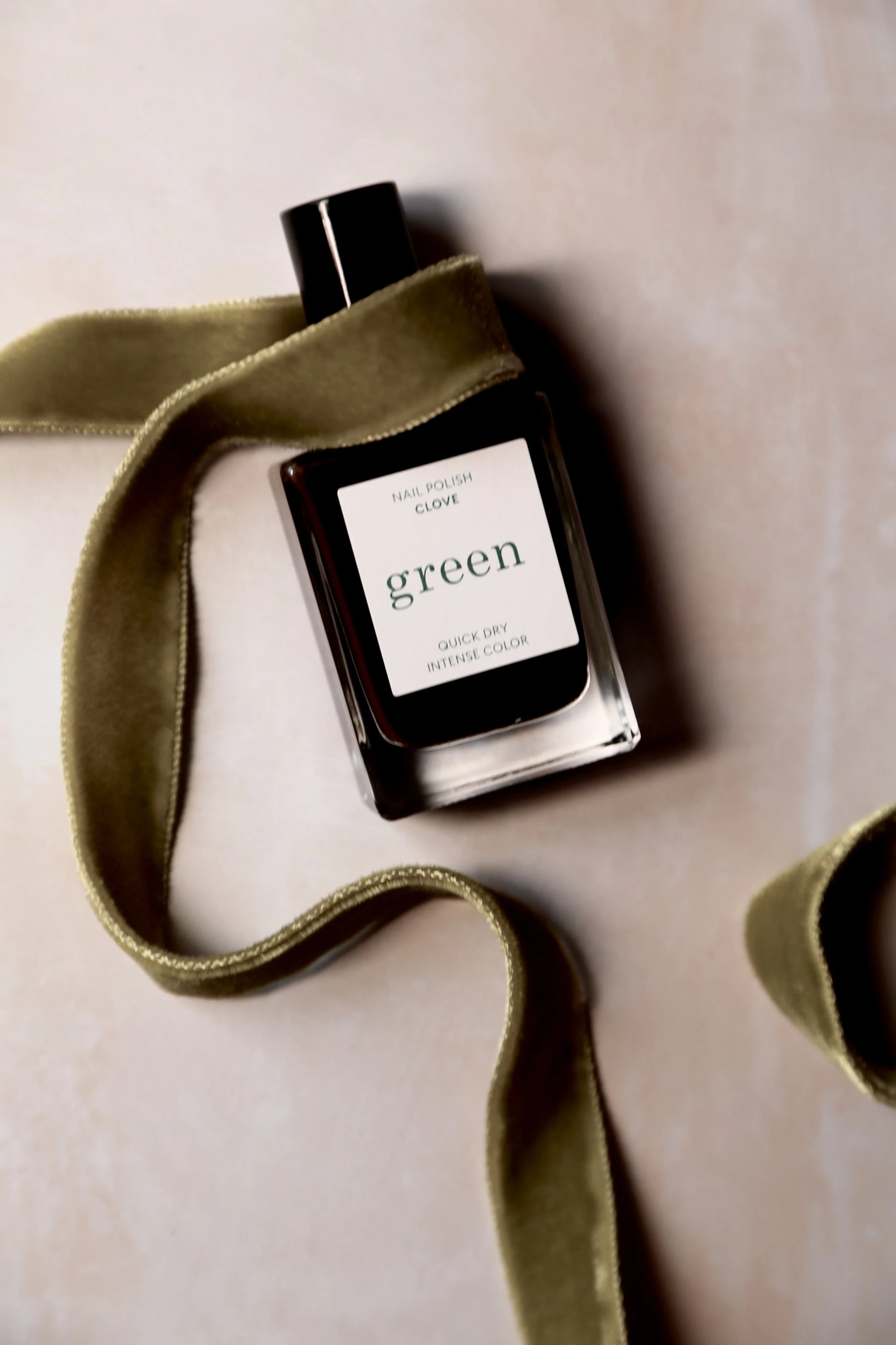 A bottle of green nail polish with a black cap, labeled 'Nail Polish Clove Green Quick Dry Intense Color,' resting on a beige surface with a green velvet ribbon wrapped around it.