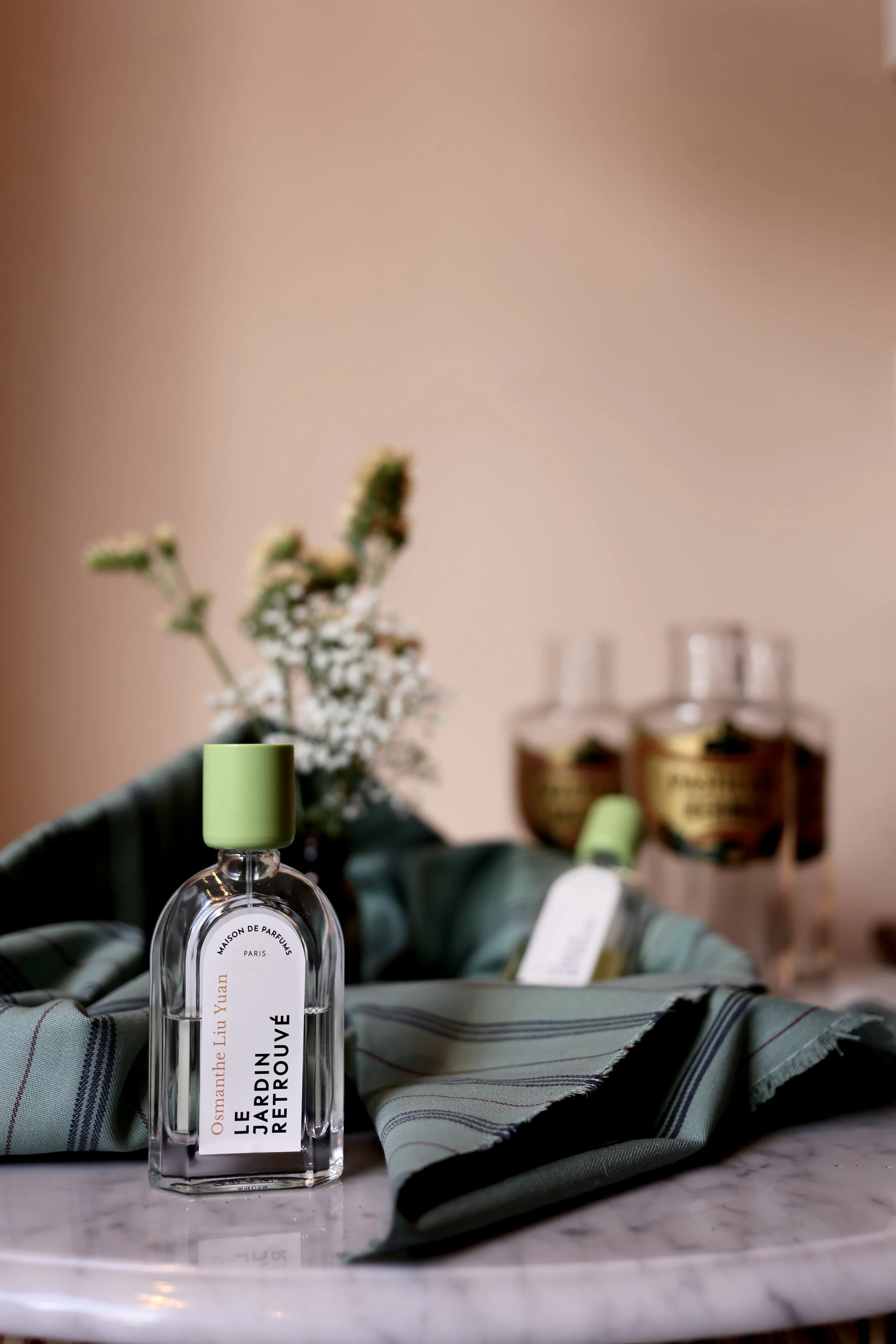 A perfume bottle with a green cap, labeled 'Le Jardin Retrouvé' by Osmanthus Liu Yuan, placed on a marble surface near a folded green and black striped cloth. In the background, a vase with white flowers and two more perfume bottles are visible.