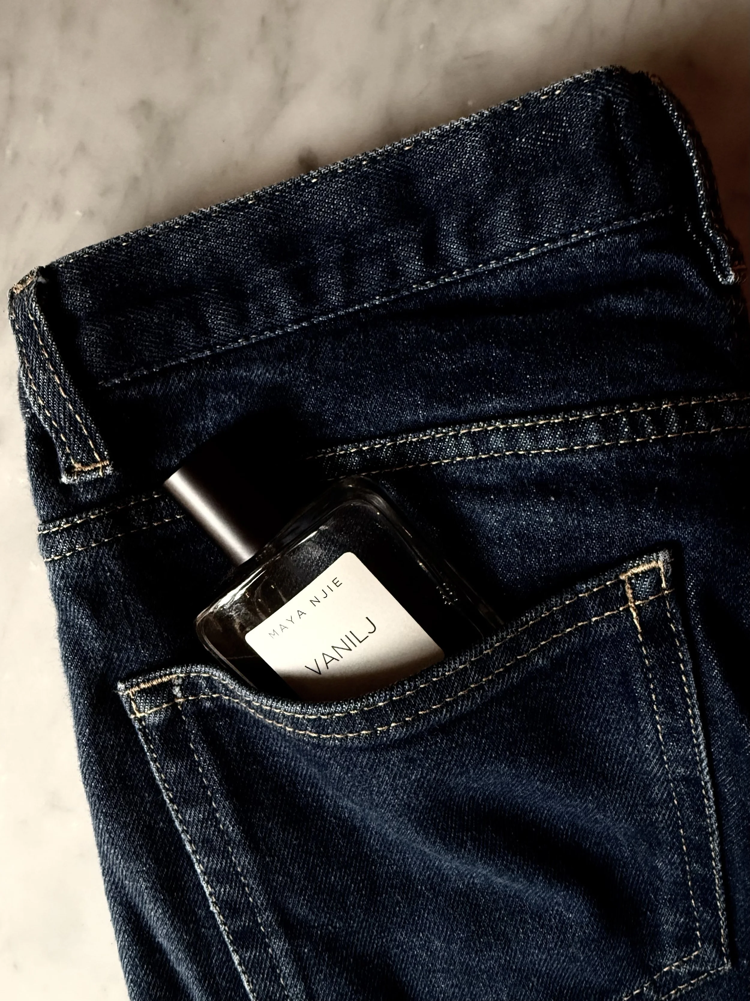 A bottle of vanilla j scented perfume partially visible in the back pocket of a pair of dark blue denim jeans.