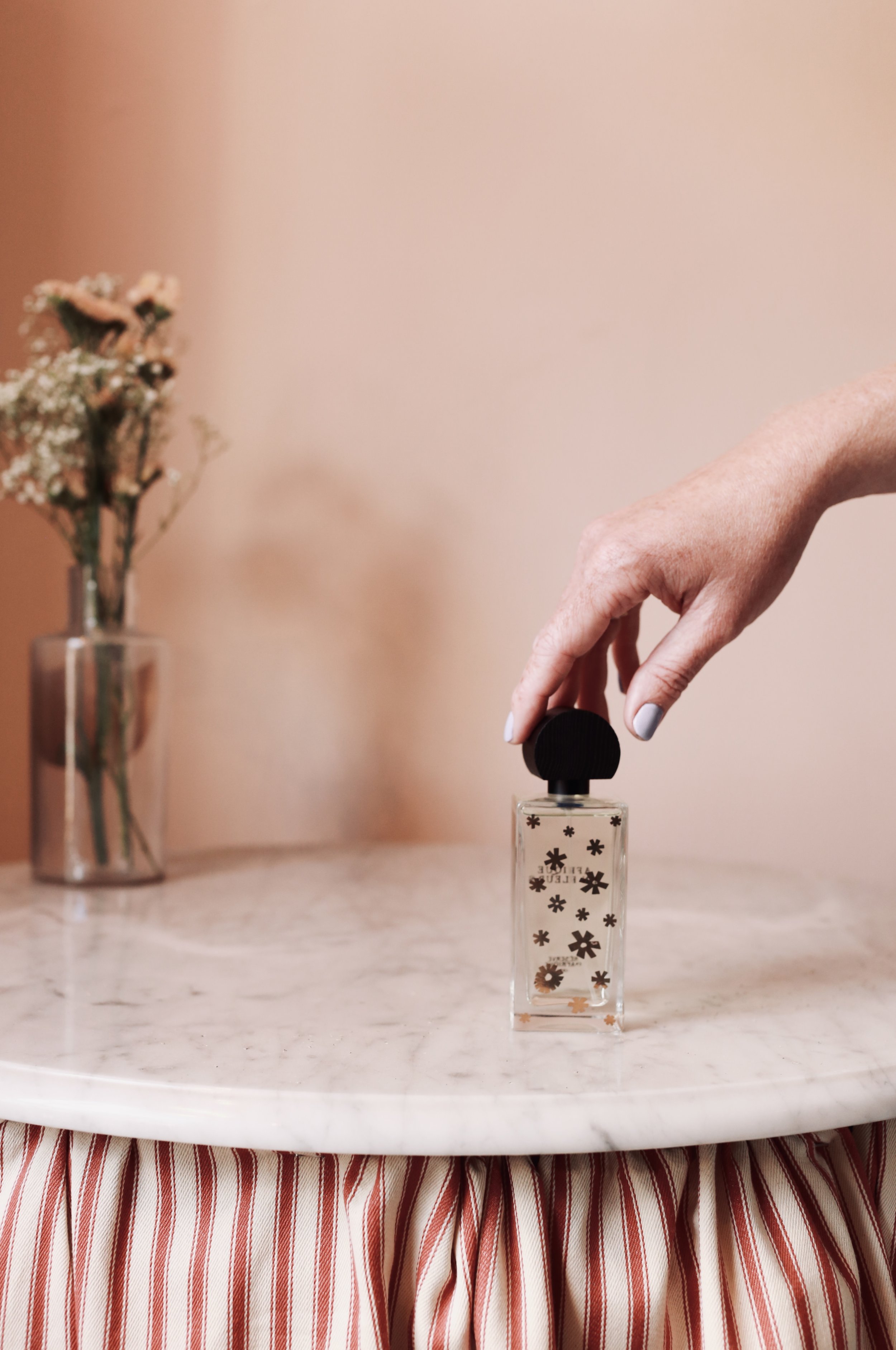 Hand reaching to pick up a bottle of perfume with black cap and floral patterns on it, on a round marble table with a striped cloth skirt, in front of a pink wall and a vase with flowers.