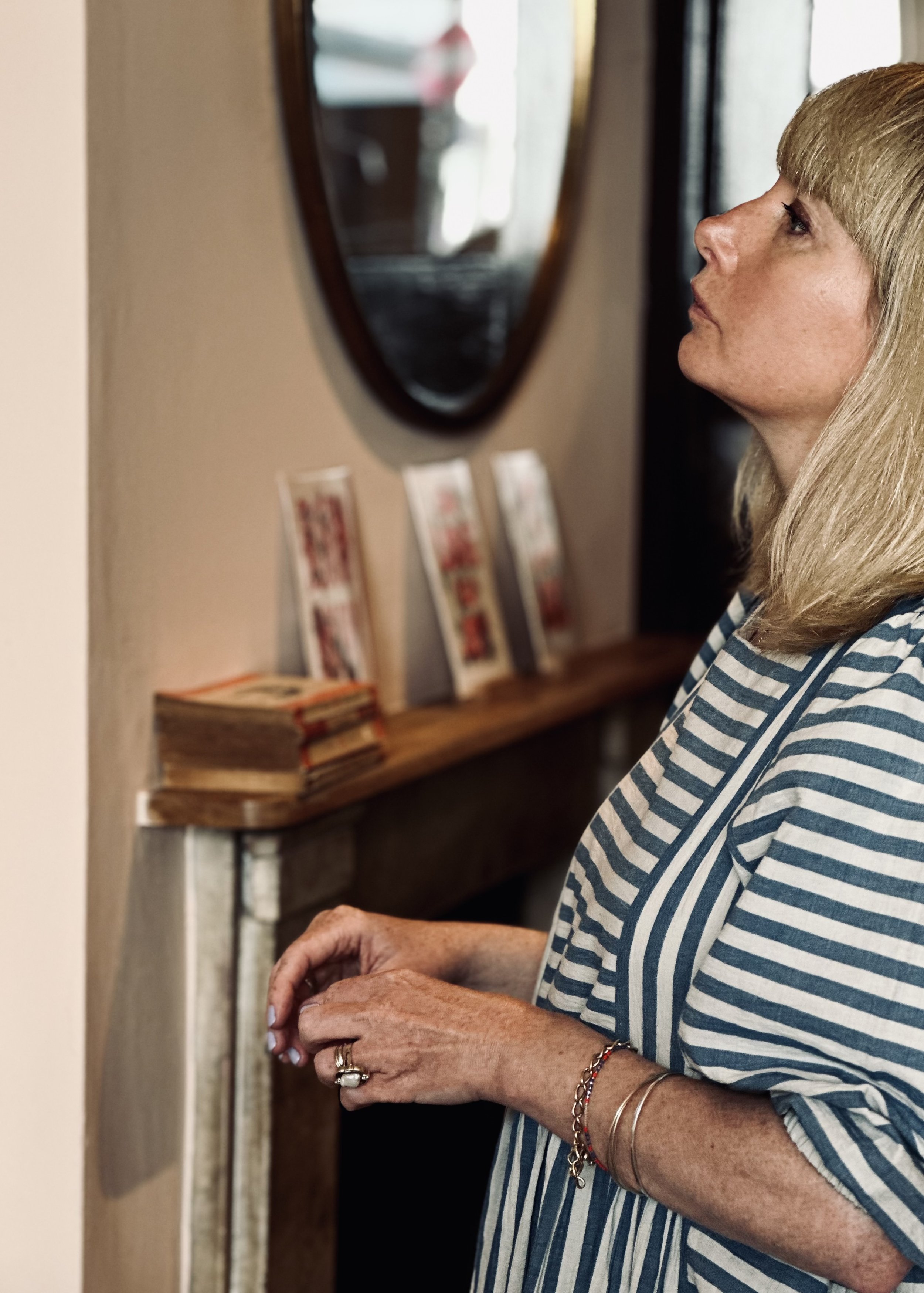 A woman with blonde hair wearing a blue and white striped shirt and jewelry, standing near a fireplace with a mirror and small framed photos on the mantle.