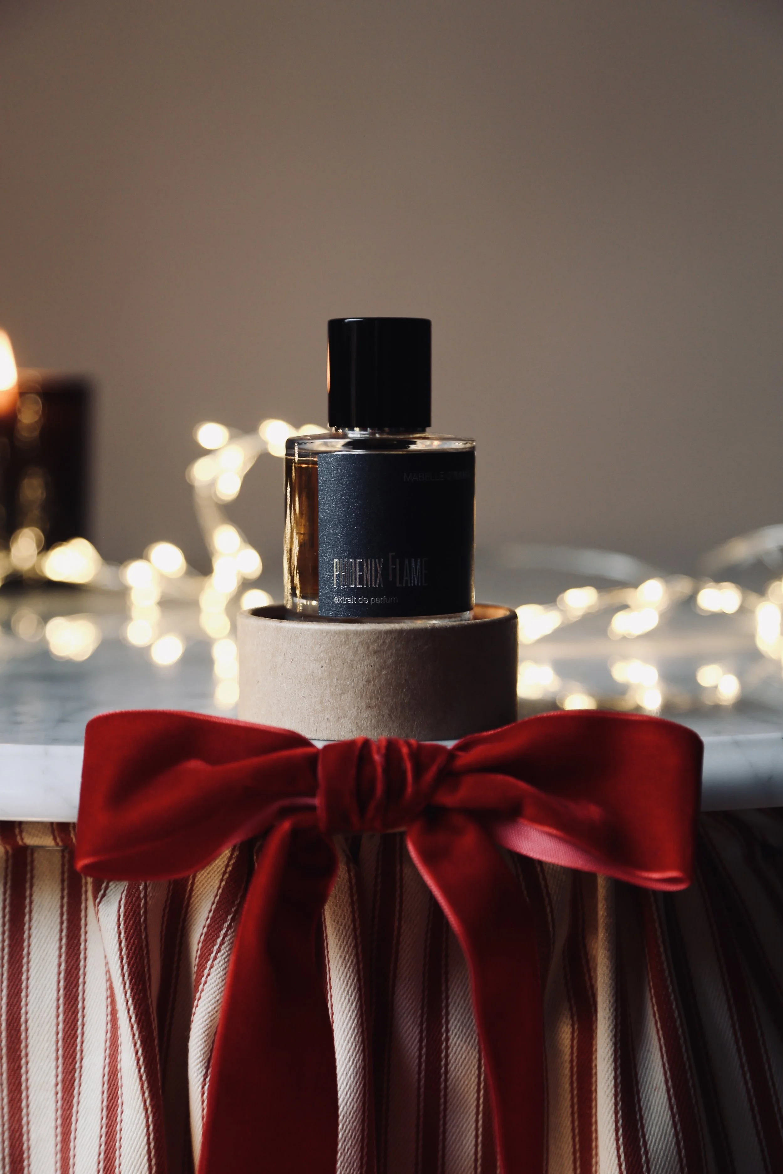 A perfume bottle labeled 'Puidenx Flame' placed on a beige cylindrical stand, with a red ribbon tied in a bow in front. The table is decorated with string lights in the background, creating a cozy, festive atmosphere.