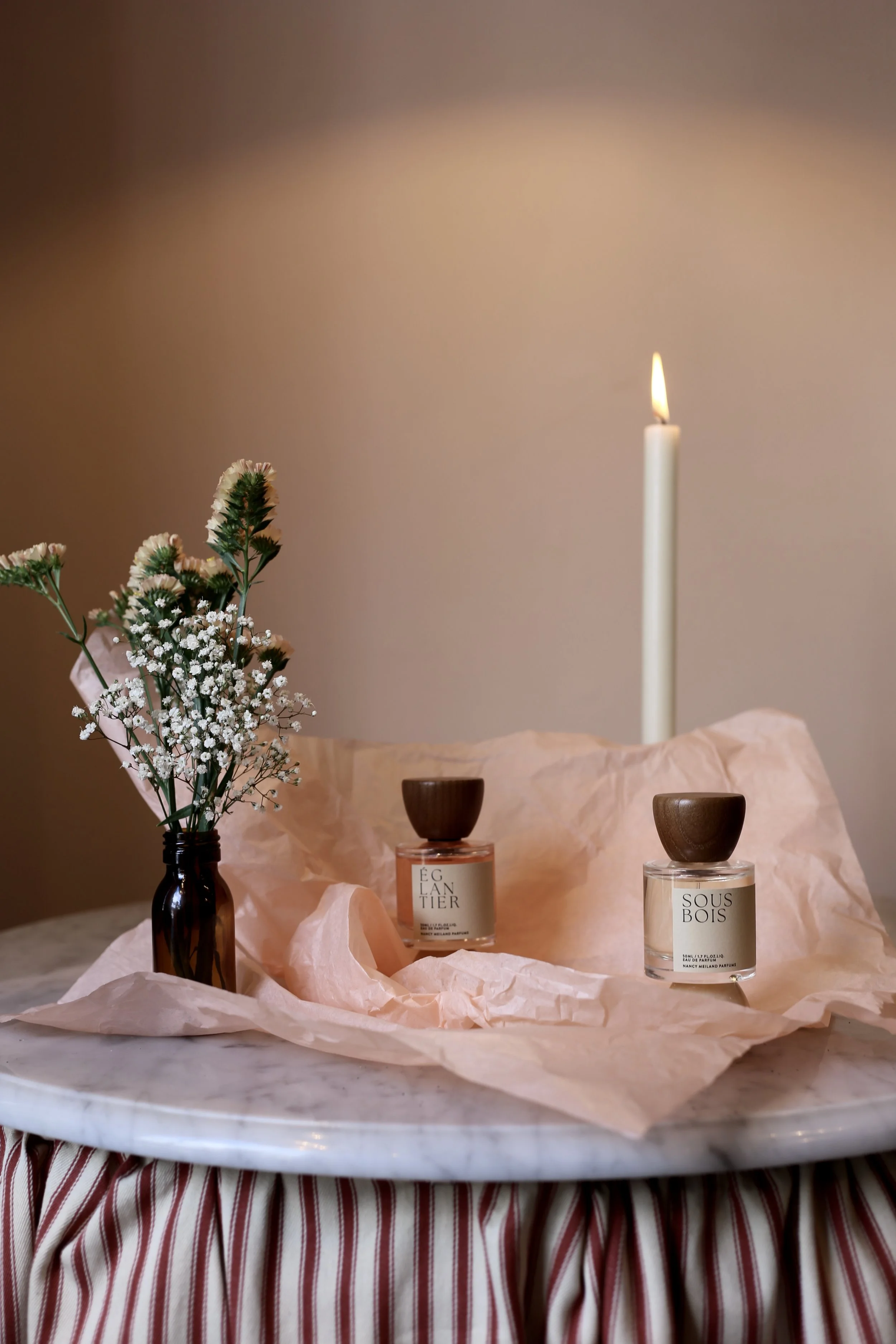 A lit candle, a small vase with flowers, and two perfume bottles on a marble table with a pink cloth, against a neutral background.
