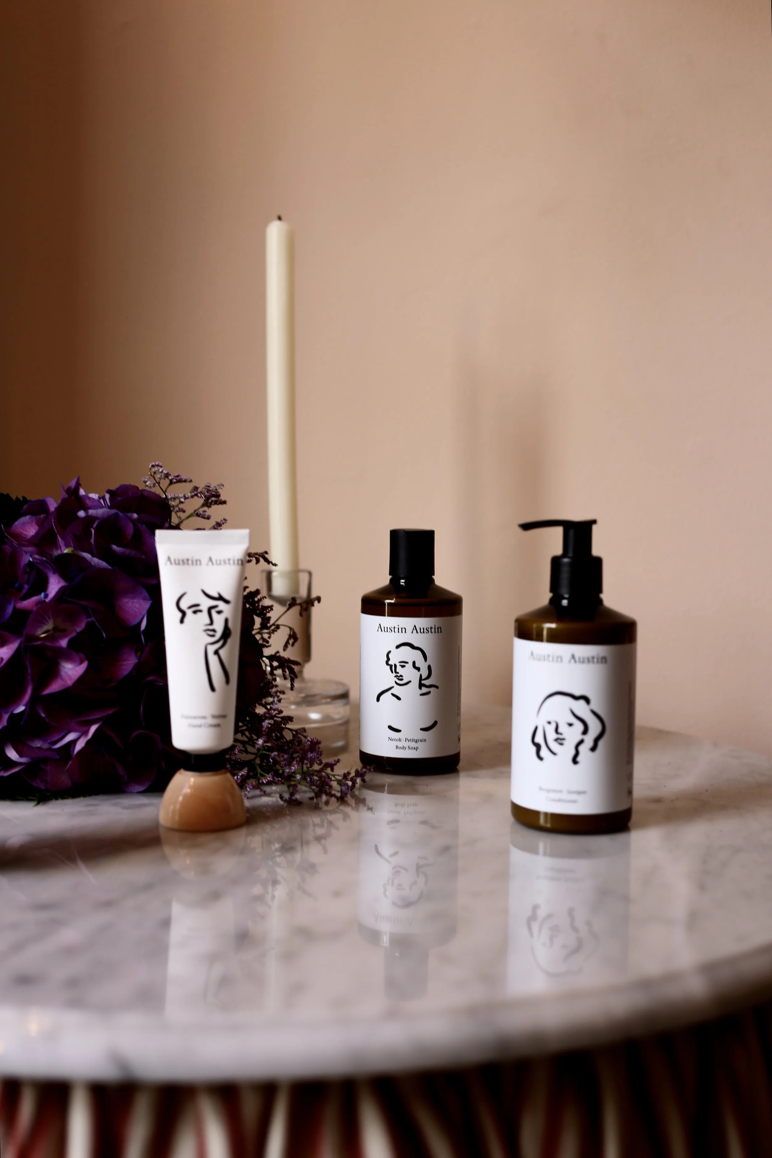 Three Austin Austin skincare bottles on a marble surface, with a purple flower arrangement and a white candle in the background.