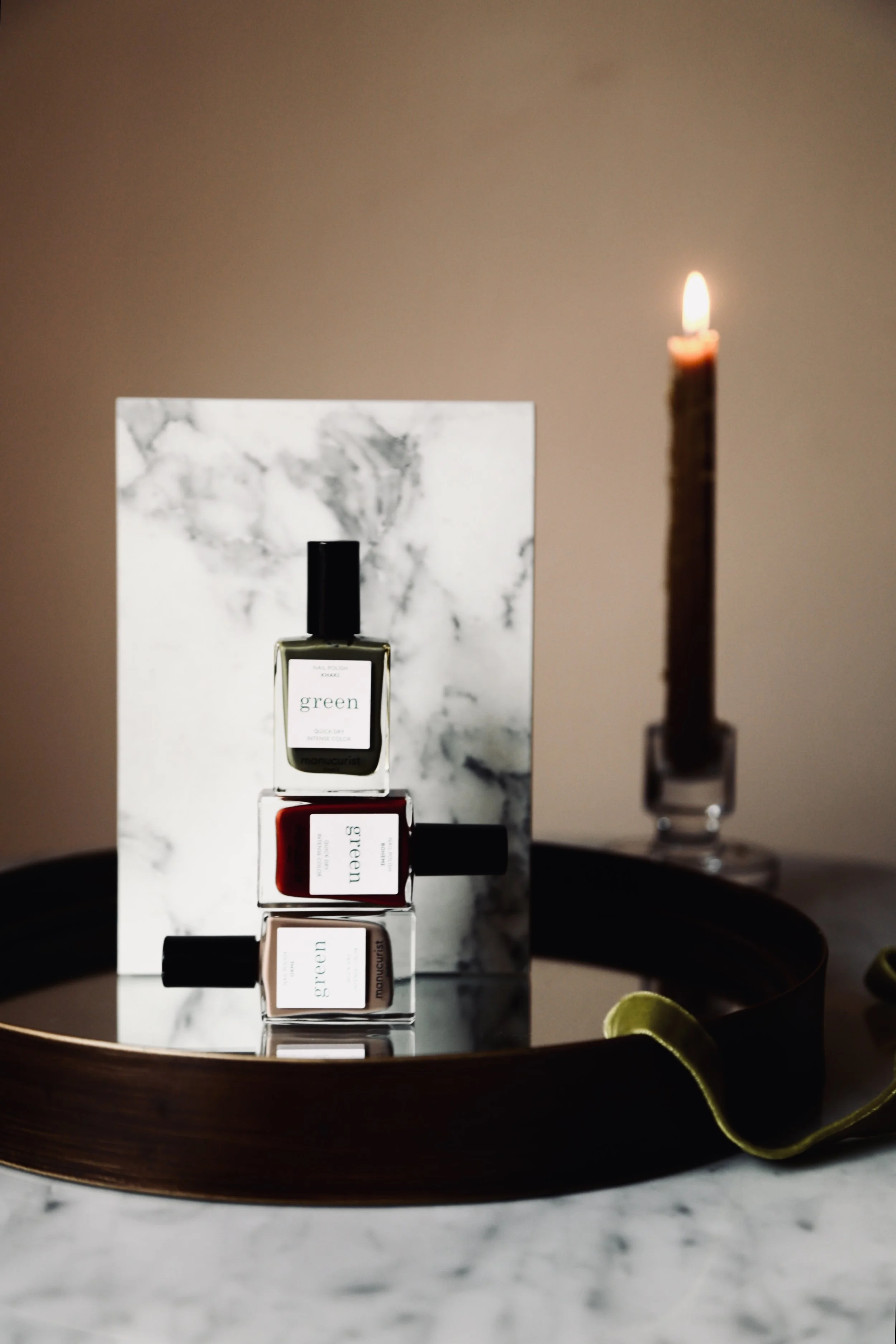 Three rectangular bottles of nail polish, stacked horizontally, on a reflective surface with a marble backboard, in front of a candle in a glass holder, all on a tray.