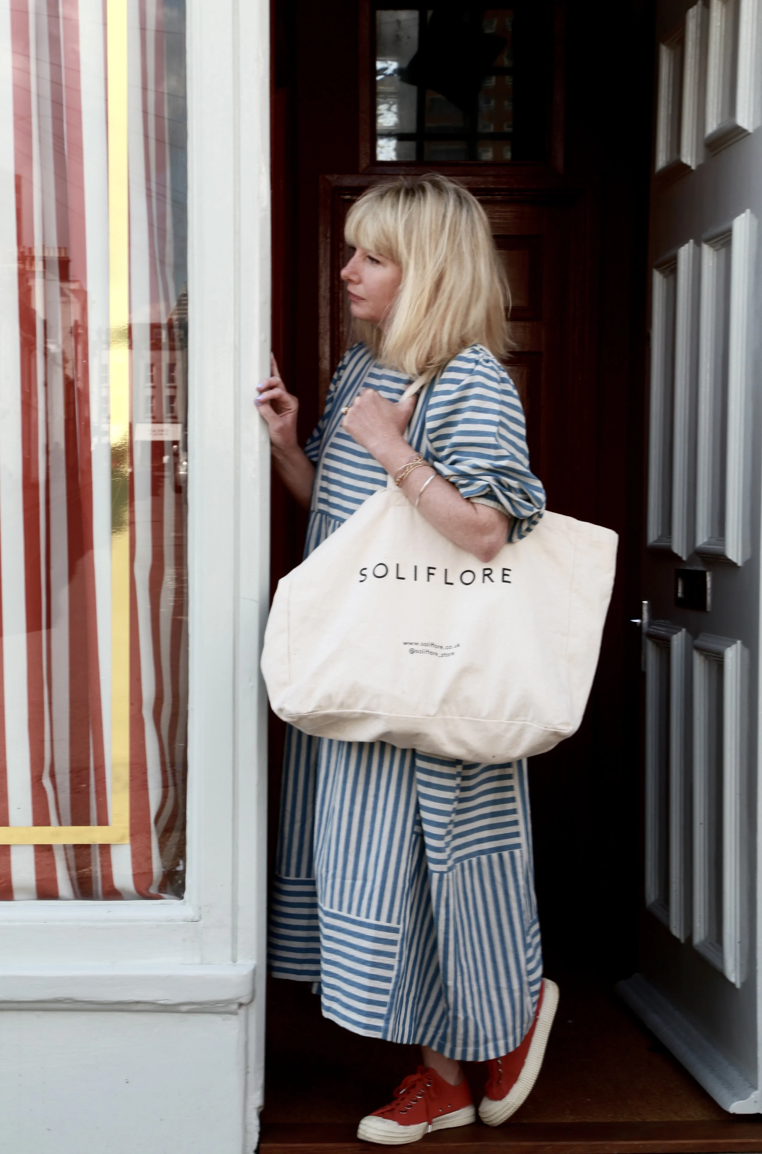 A woman with blonde hair is standing at a doorway, holding a tote bag with 'SOLIFLORA' printed on it. She is wearing a blue and white striped dress and red sneakers.