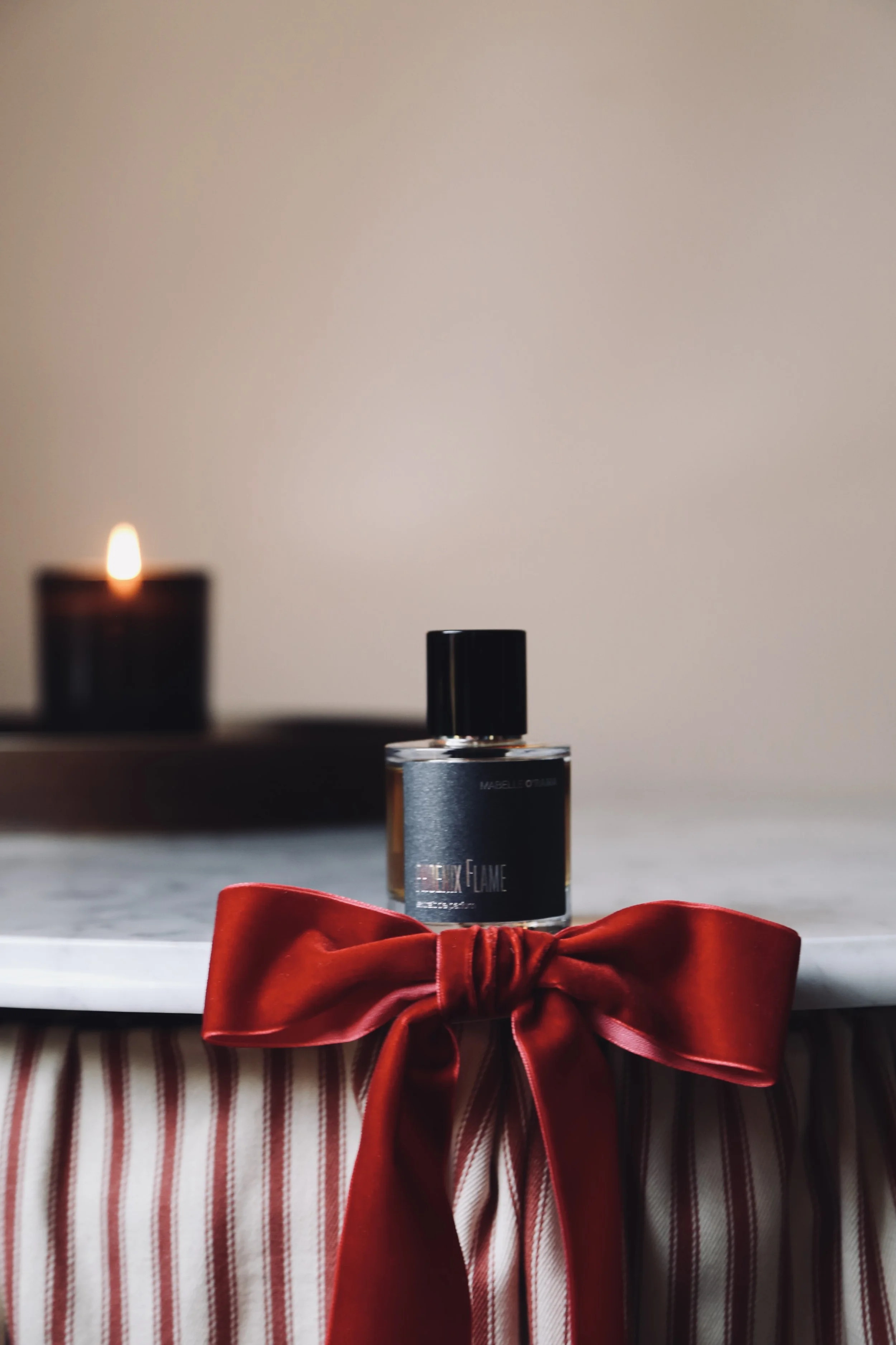 A perfume bottle labeled 'Flick Flame' sits on a white marble surface decorated with a red satin bow. In the background, a lit candle is visible.