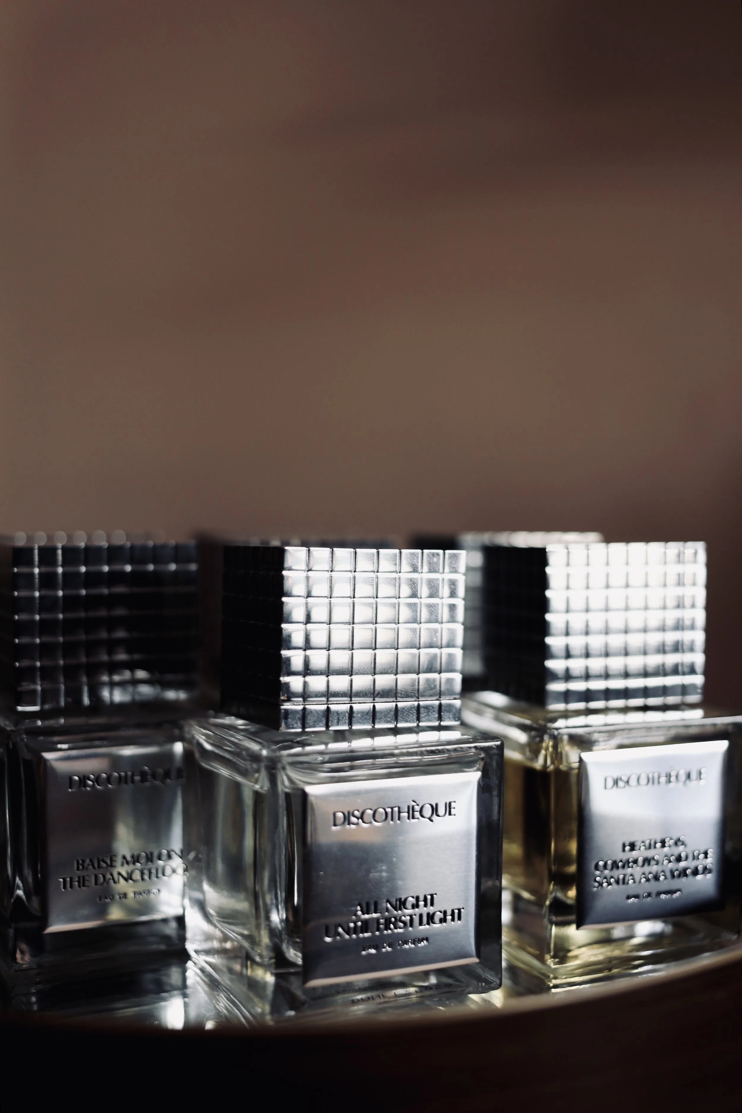 Three bottles of perfume with metallic cube caps, placed on a reflective surface. The bottles have silver labels with the brand name 'Discohèque' and fragrance names such as 'All Night' and 'Hearts.'
