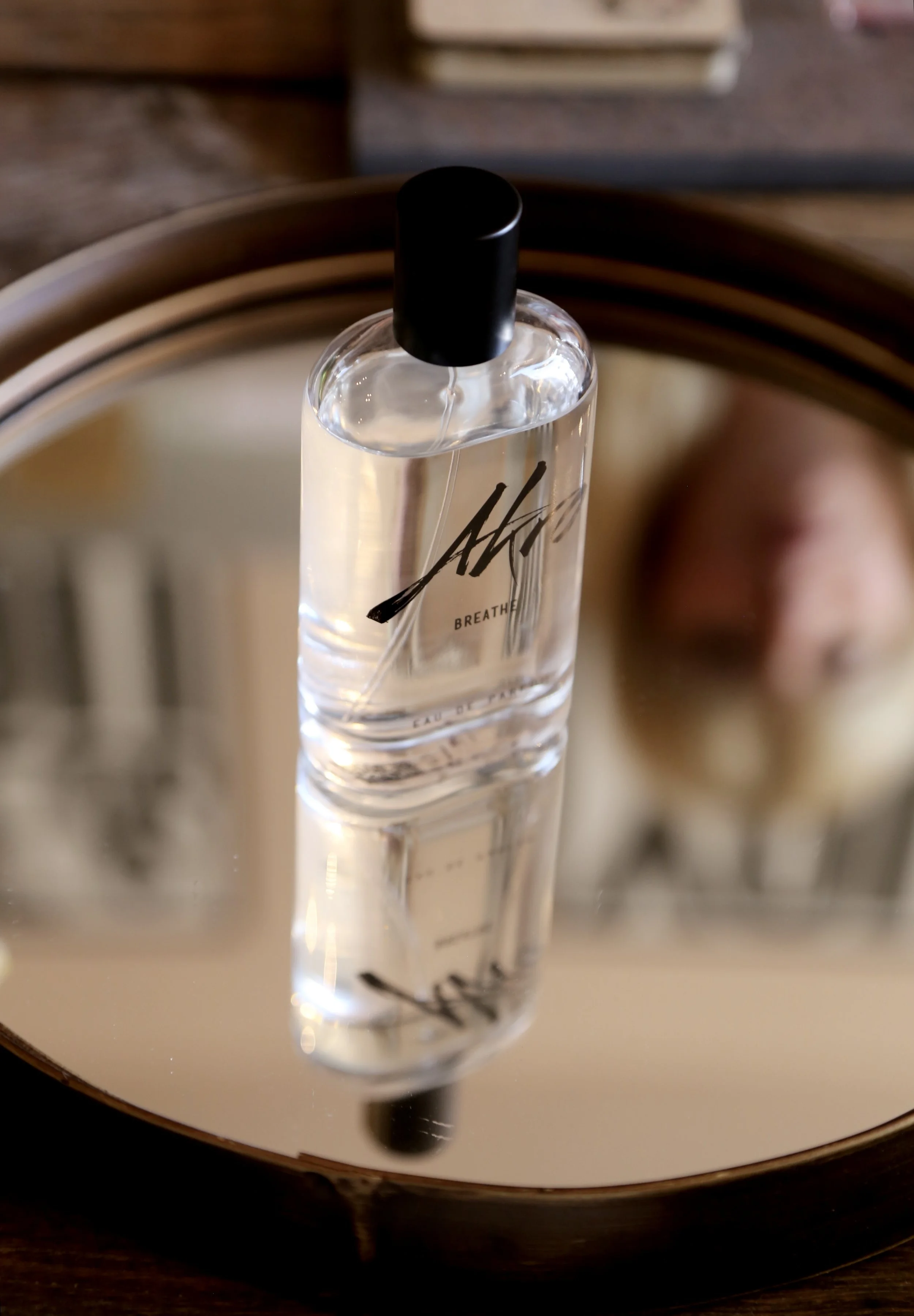 A clear perfume bottle with black cap and black abstract signature lettering, placed on a round mirrored tray with a wooden edge.