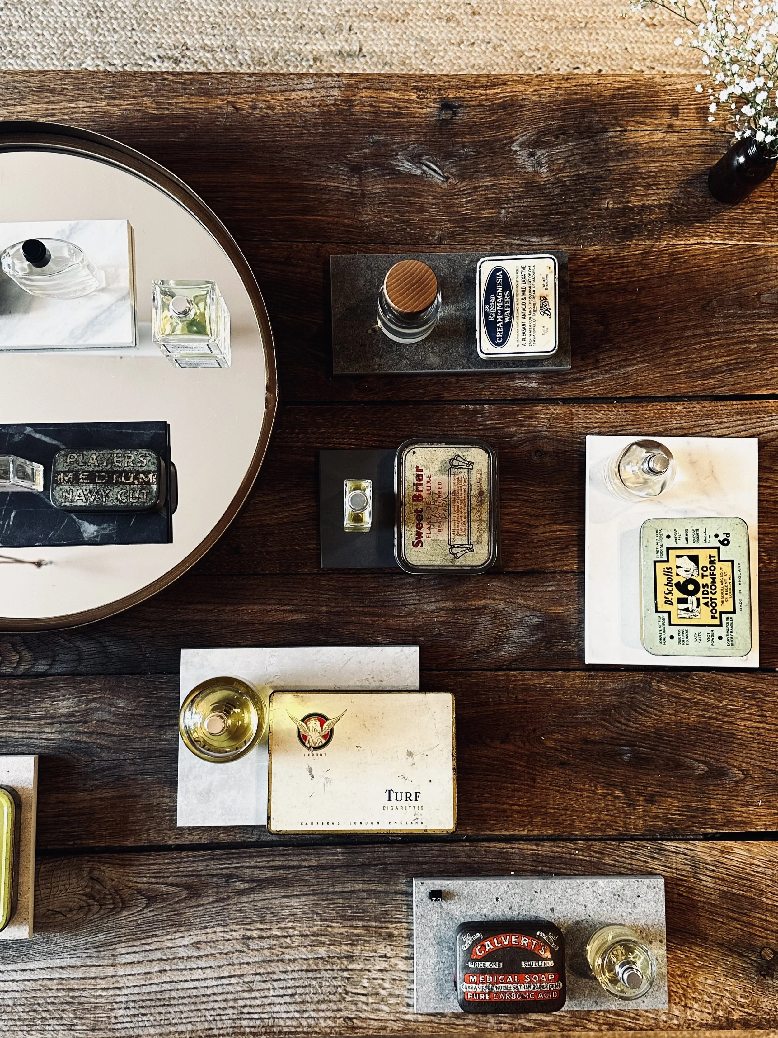 Top-down view of a dark wooden table with various vintage items including cigarette packs, a glass of white wine, perfume bottles, and small decorative objects.