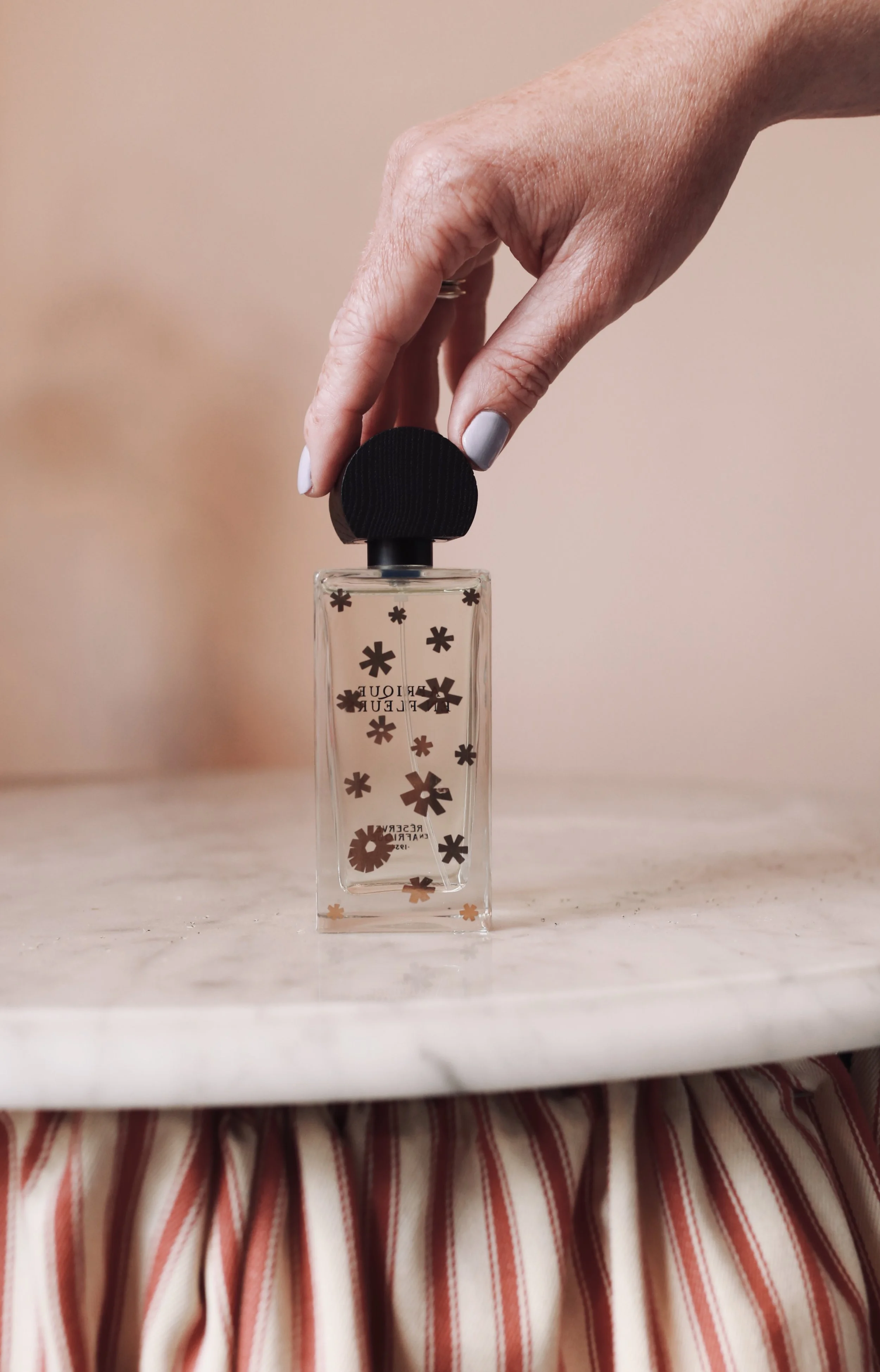 A hand with light skin tone and painted nails holding a black cap of a clear rectangular perfume bottle with black flower patterns on a light-colored surface.
