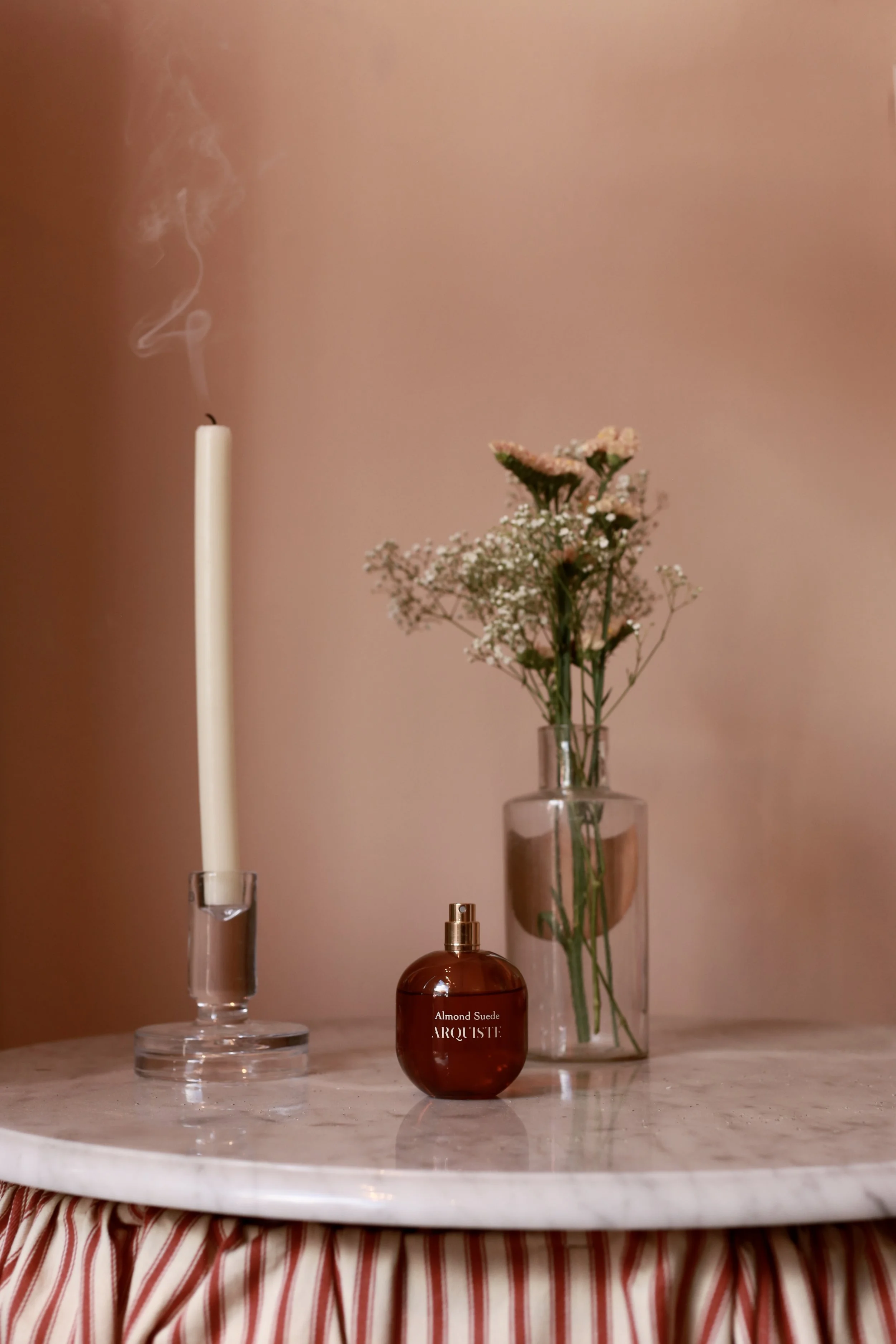 A lit white candle with smoke rising, a small bouquet of pink and white flowers in a clear glass vase, and a round amber perfume bottle labeled 'Almond Suede' on a marble surface with a pink wall background.