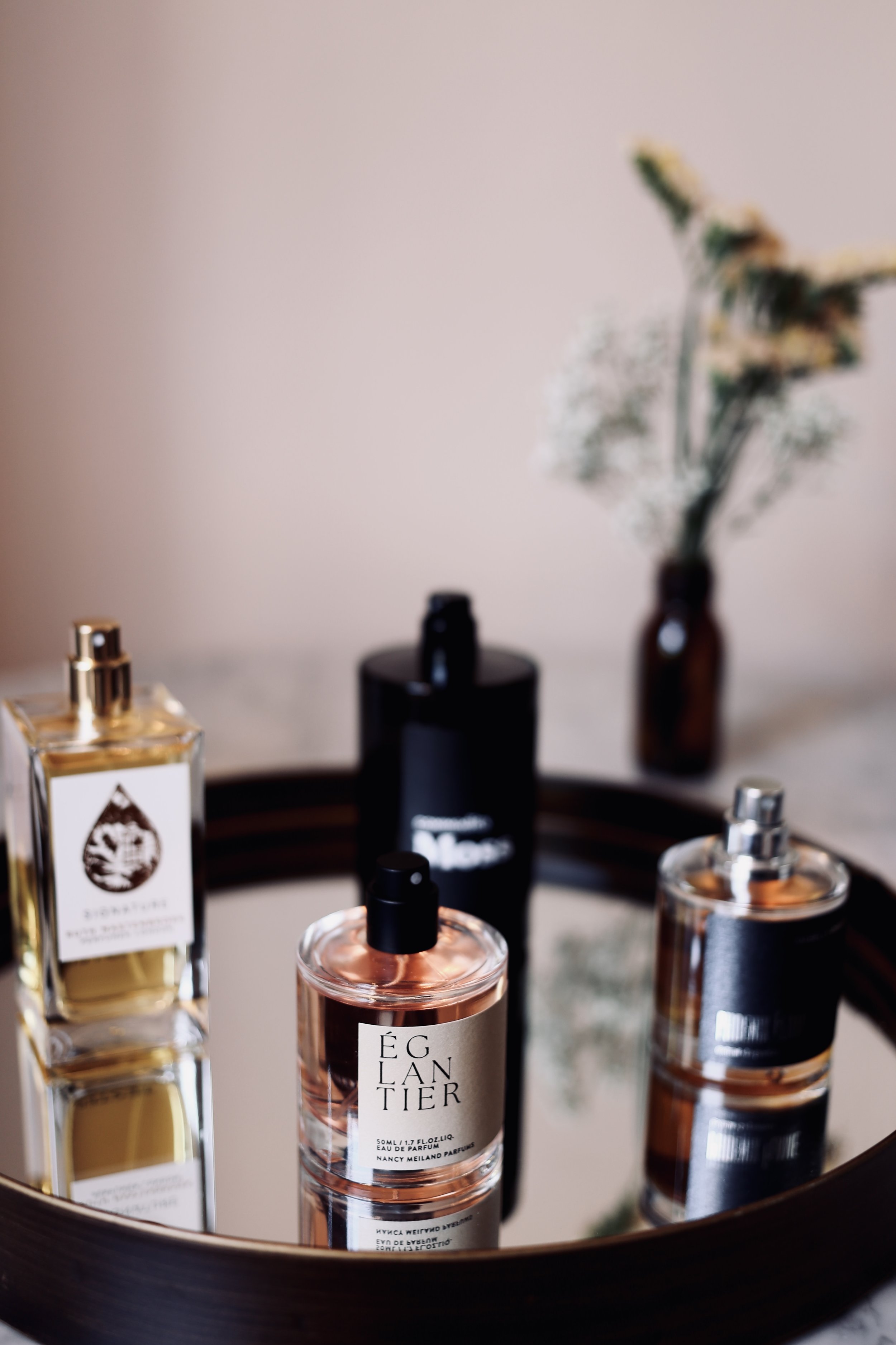 A round mirror tray holding perfume bottles, with a vase of flowers in the background.