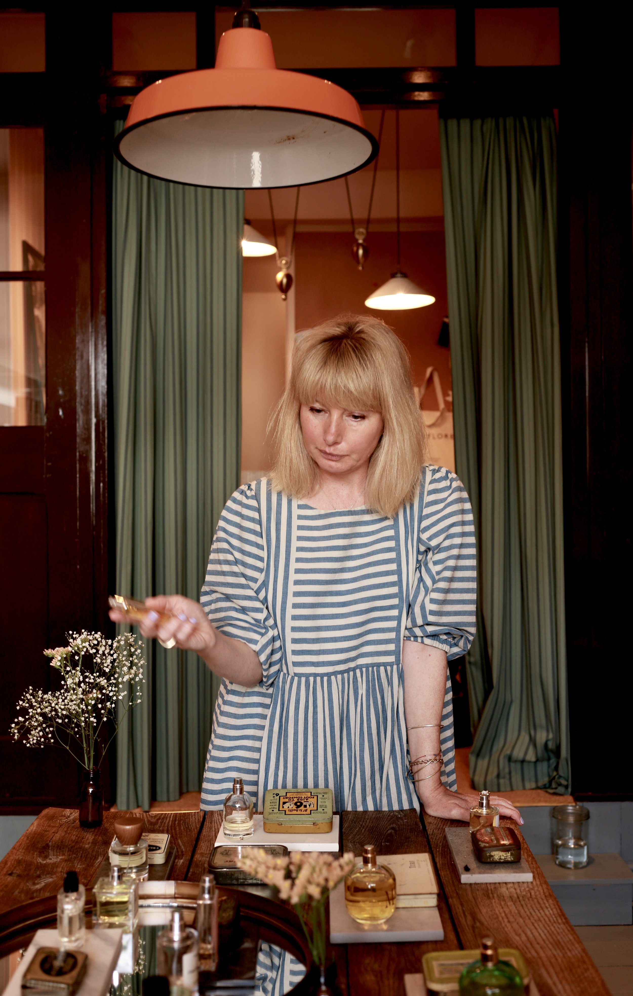 A woman with blonde hair and a striped dress standing at a table with perfumes and flowers in a cozy indoor setting with green curtains and warm lighting.