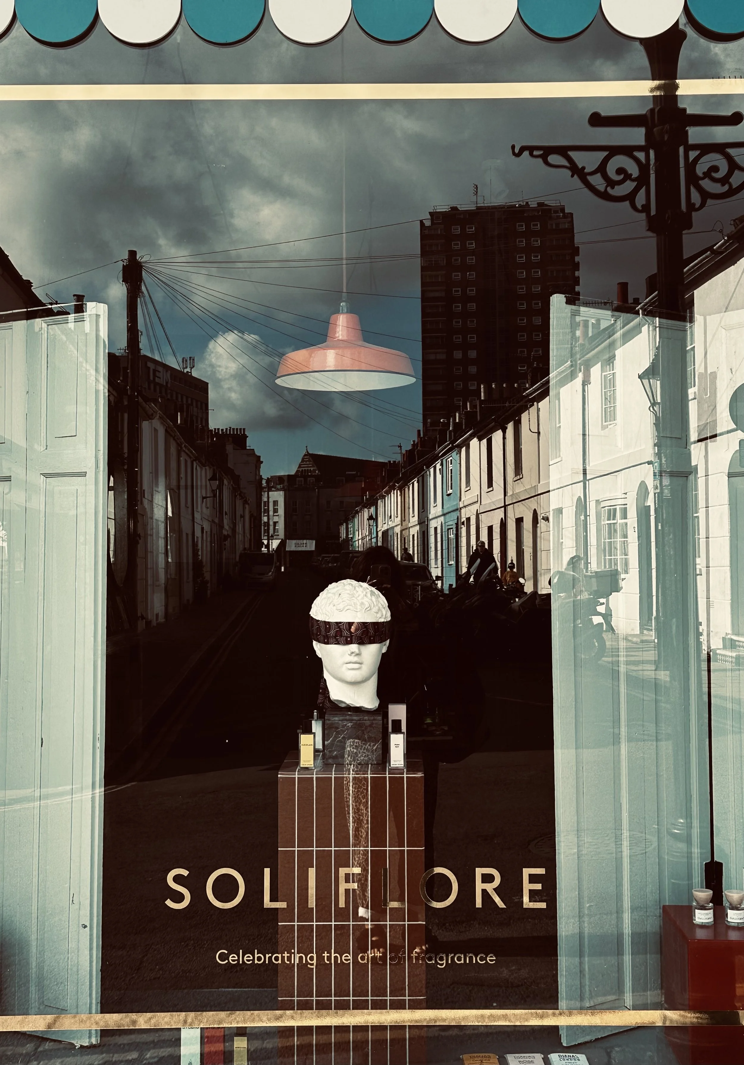 Storefront window display for Soliflore, celebrating the art of fragrance, featuring a mannequin head with a blindfold and perfume bottles, reflections of buildings and street scene outside.