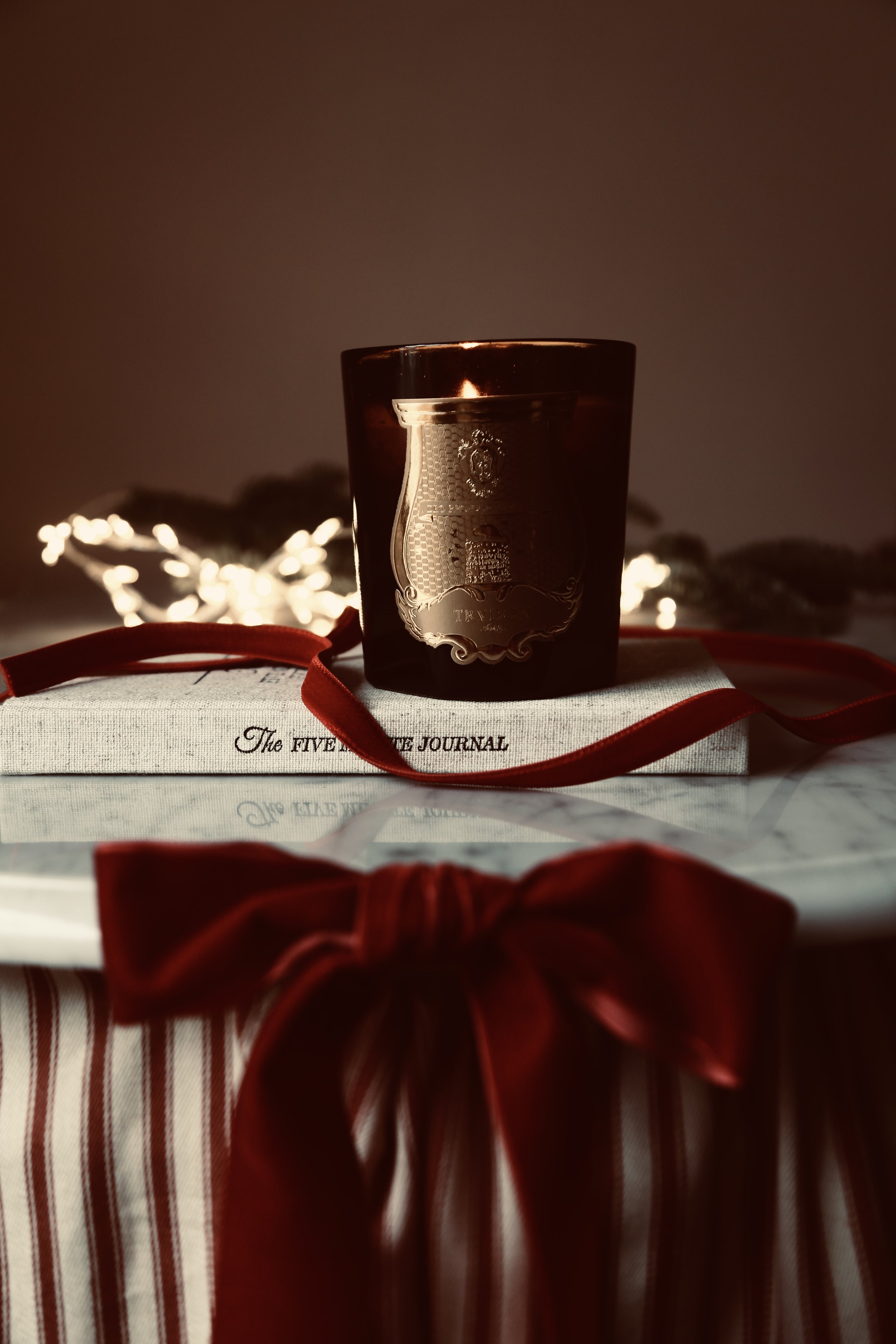 A dark brown candle in a glass holder, placed on a wrapped gift box with red ribbon, in front of a book titled 'The Five Love Languages Journal' and blurred fairy lights in the background.