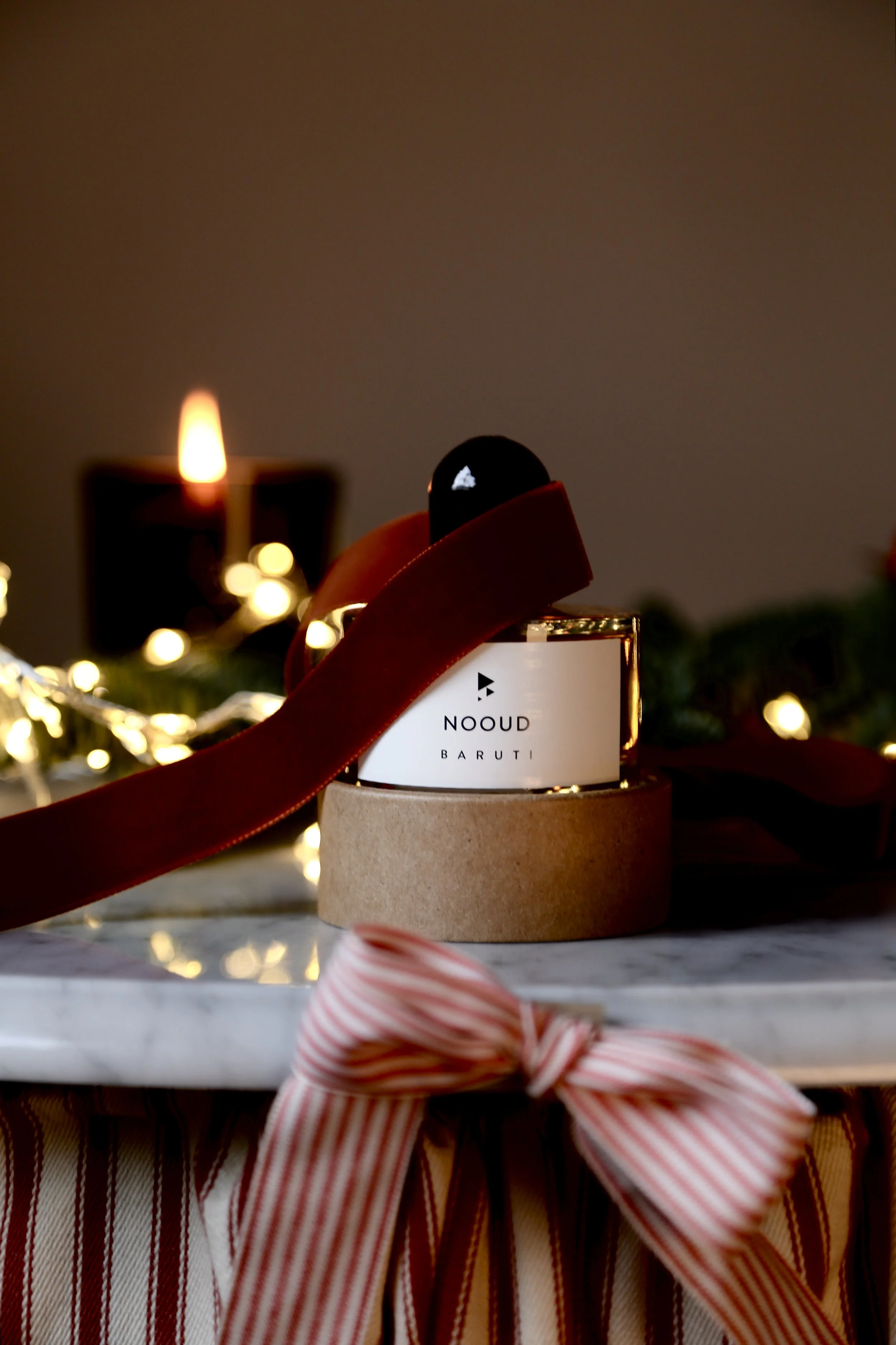 A holiday-themed display of a perfume bottle, decorated with a red velvet ribbon, placed on a marble surface with a striped cloth and festive lights in the background.