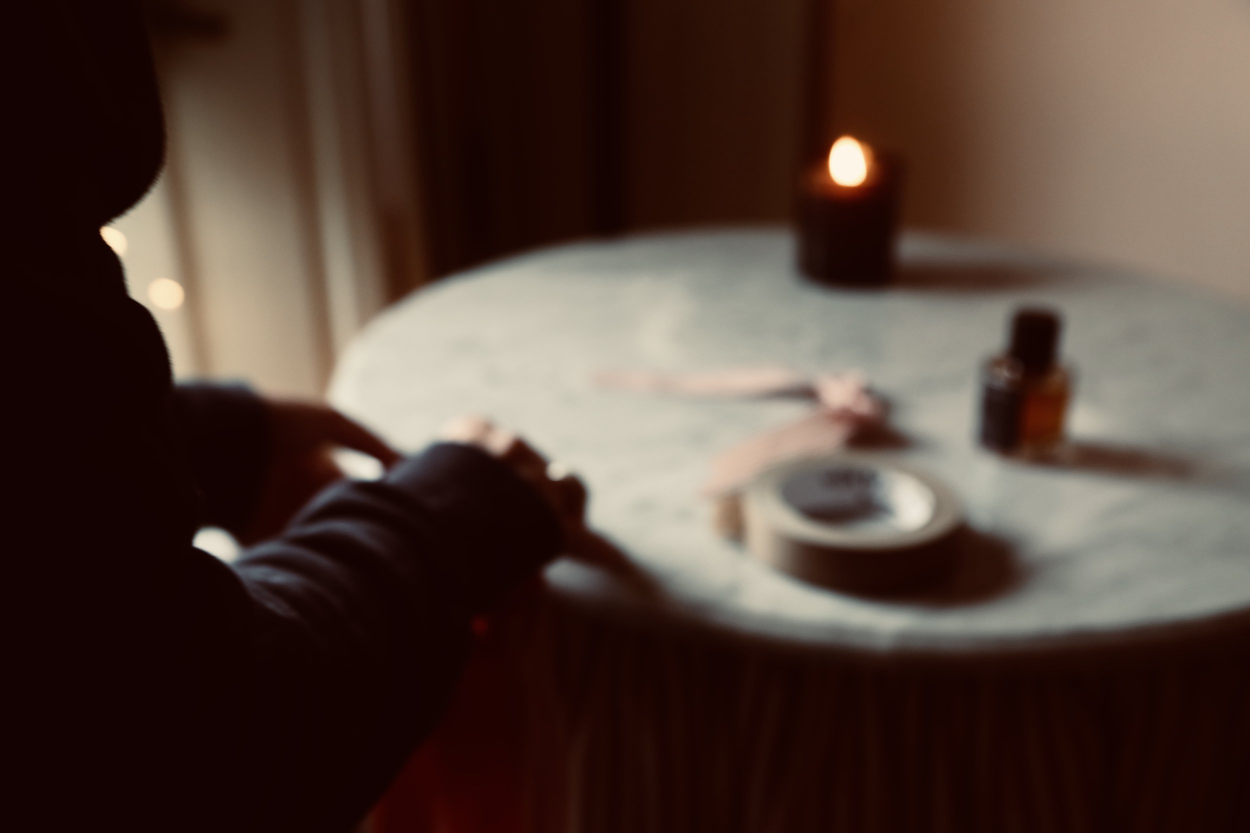 Silhouette of a person with long hair sitting at a table with a lit candle, a bottle of nail polish, and some tape on it, in a dimly lit room.