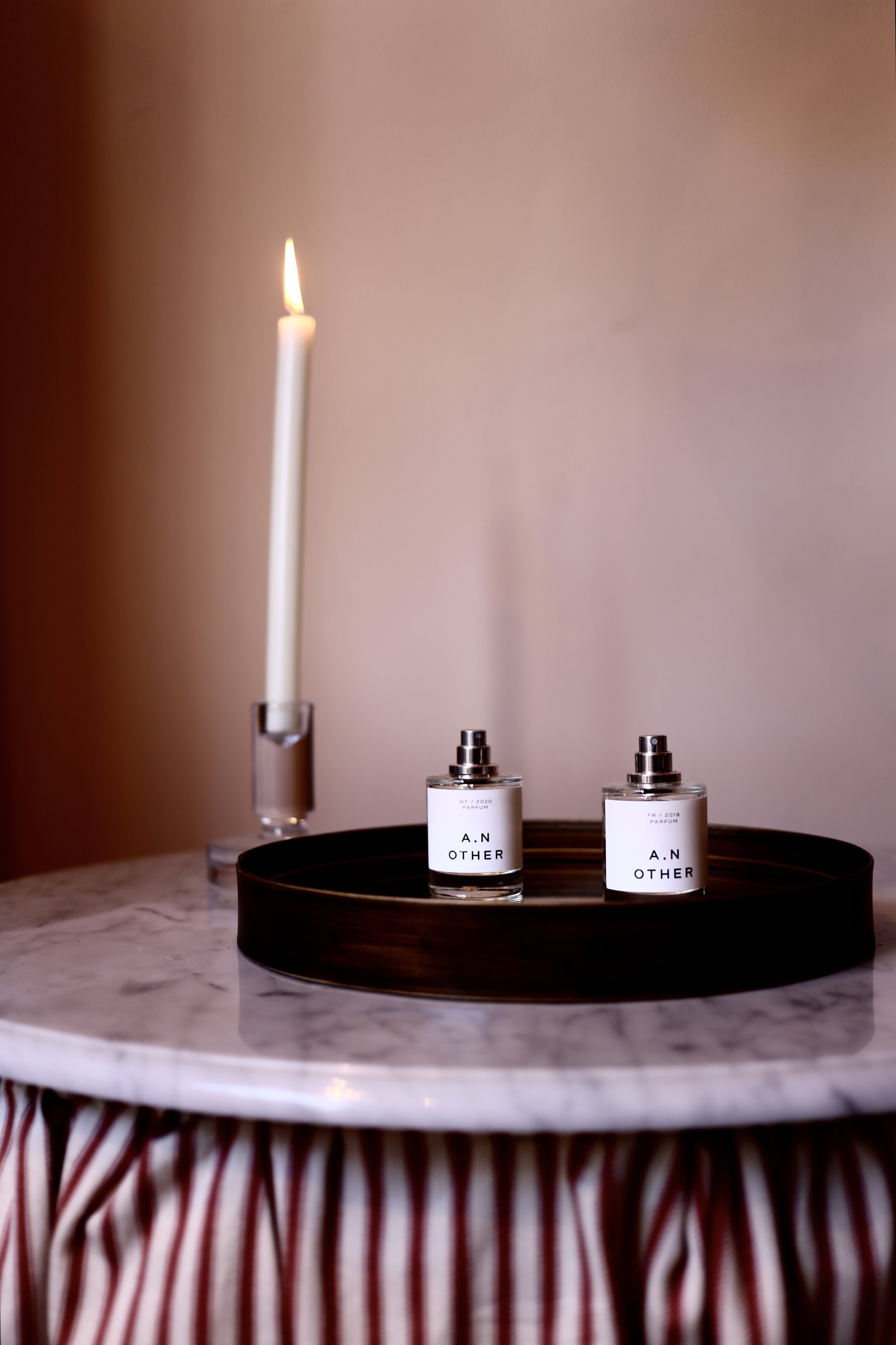 Two bottles of A.N Other perfume on a black tray, with a lit candle in a clear glass holder, placed on a marble-topped table with a red and white striped skirt.
