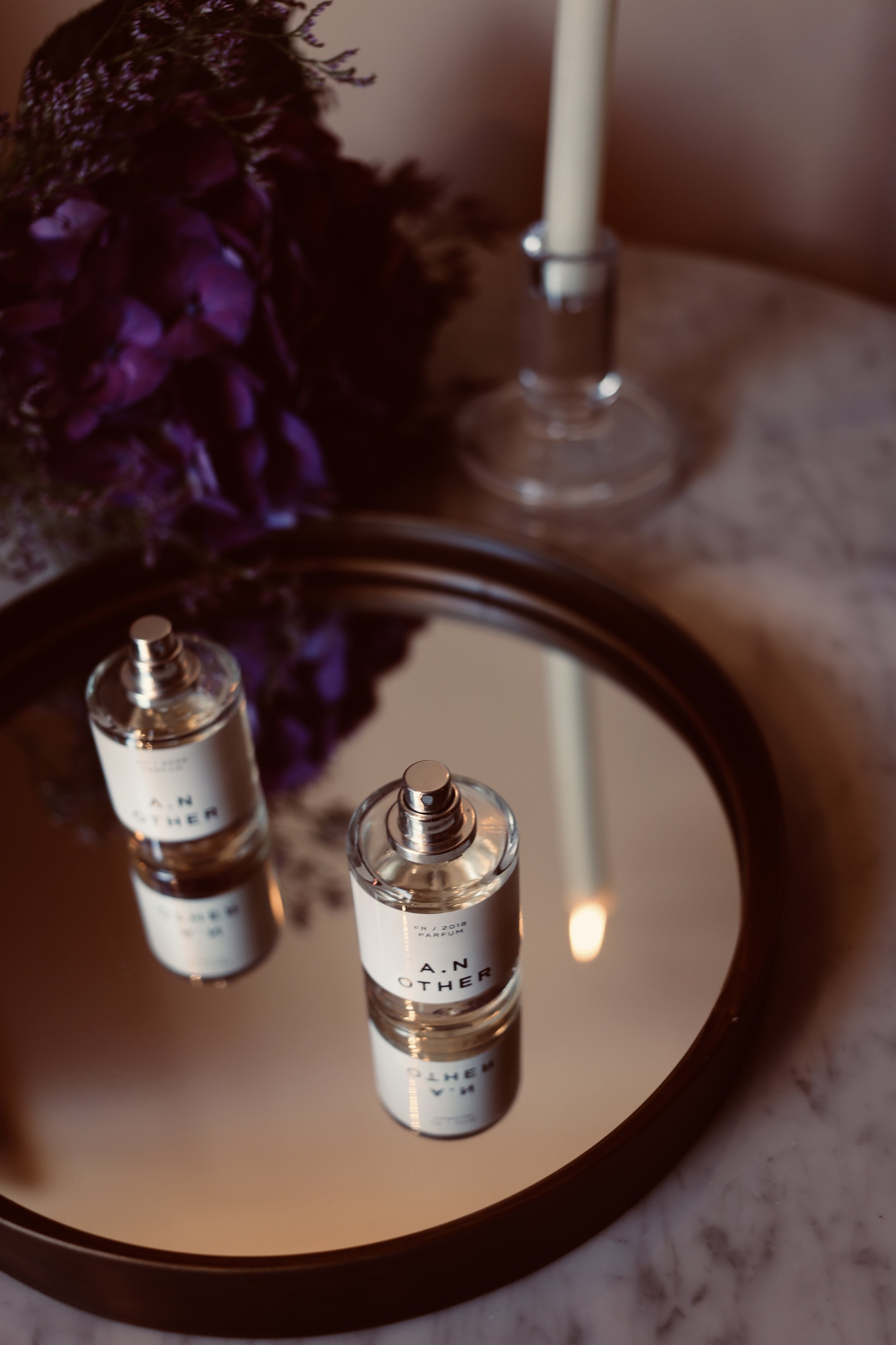 A perfume bottle labeled 'A.N.OTHER' is reflected in a mirror on a marble surface, with purple flowers and a candle in the background.