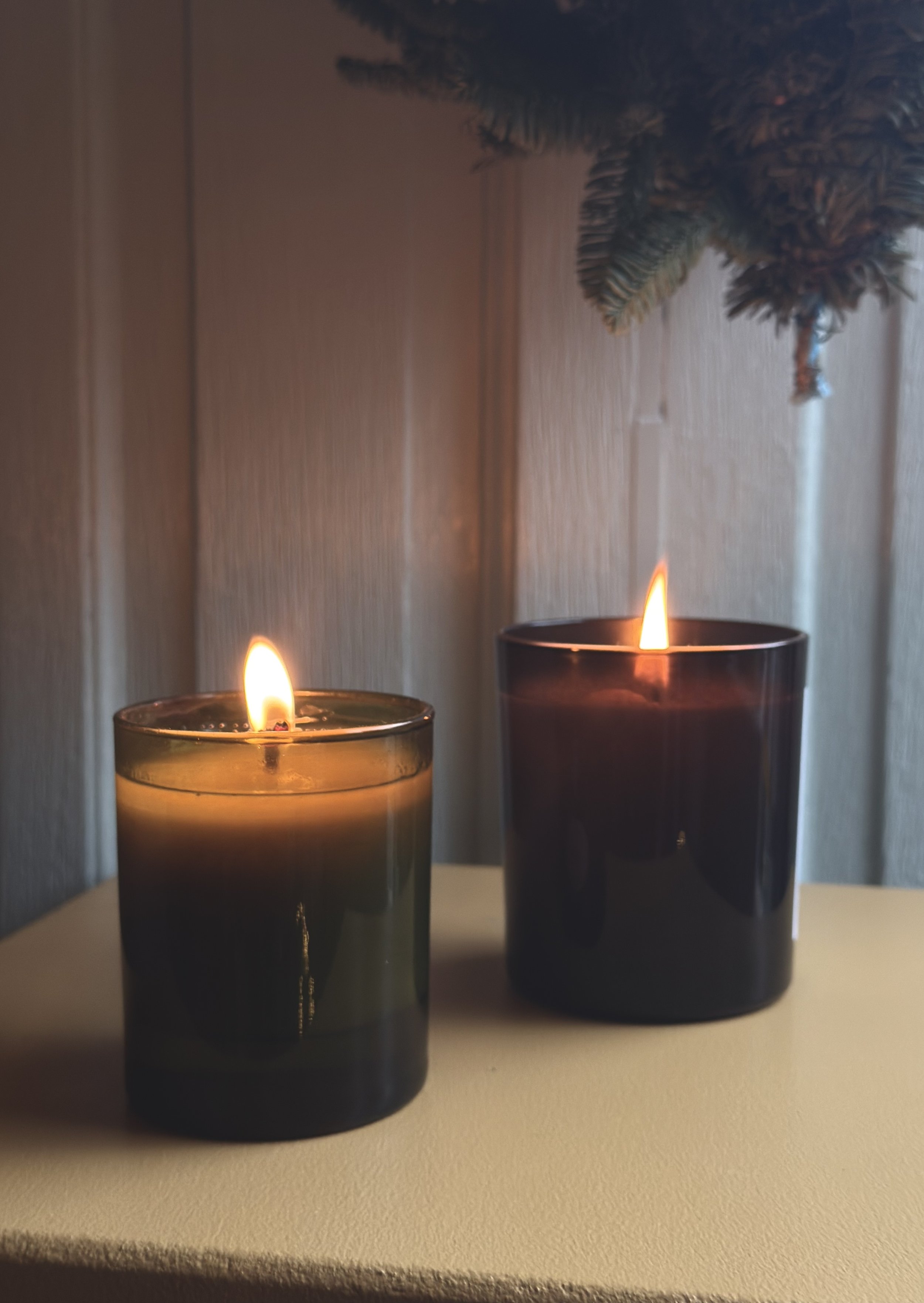 Two lit candles in glass holders placed on a light-colored surface with a wooden wall and part of a pine branch in the background.