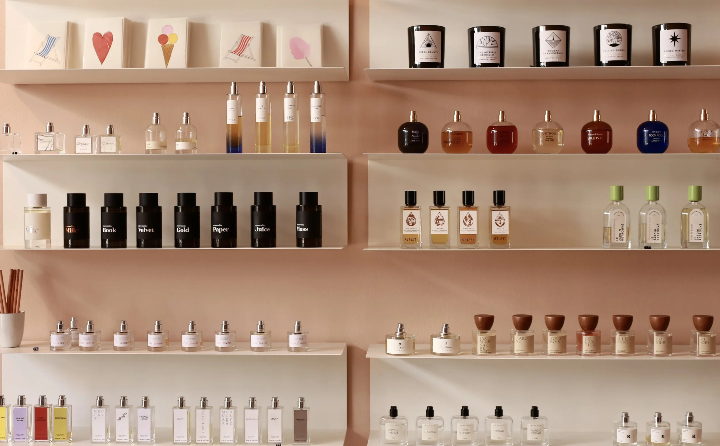 Shelves filled with perfume bottles and candles, some decorated with colorful artwork, set against a pink wall in a boutique
