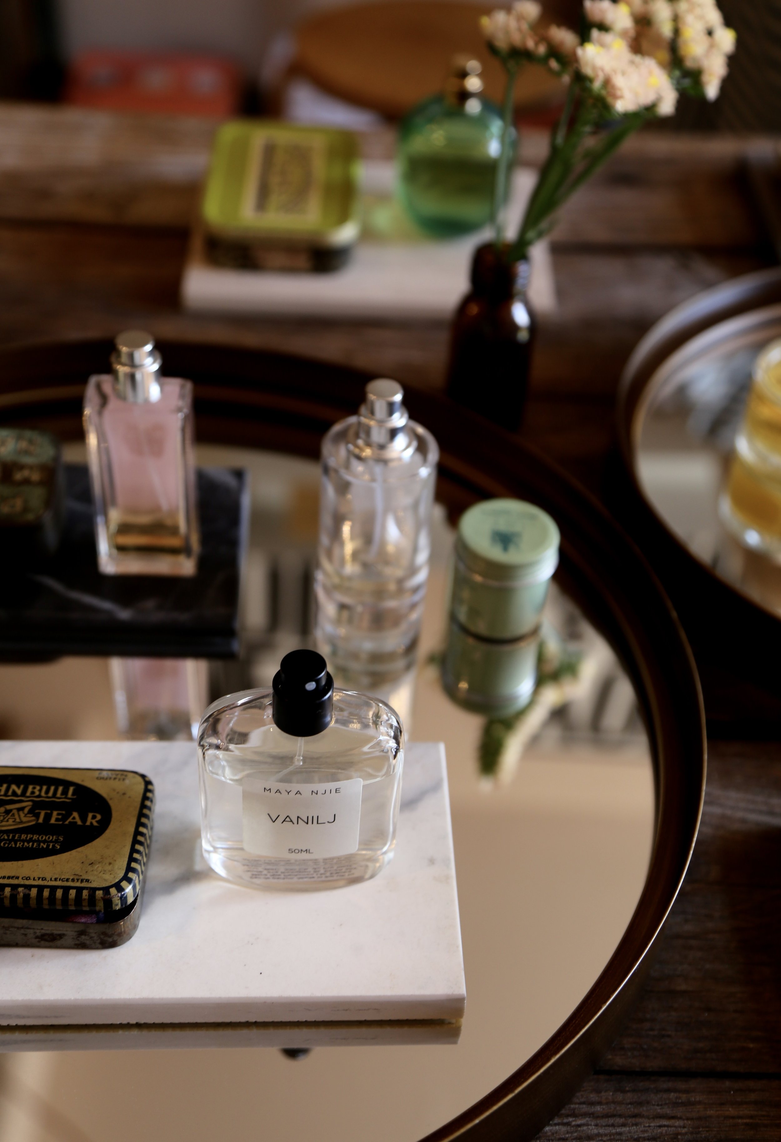 A collection of perfume bottles and cosmetics on a glass table with a white marble slab in the foreground, a small tin container, and a vase with white and pink flowers in the background.