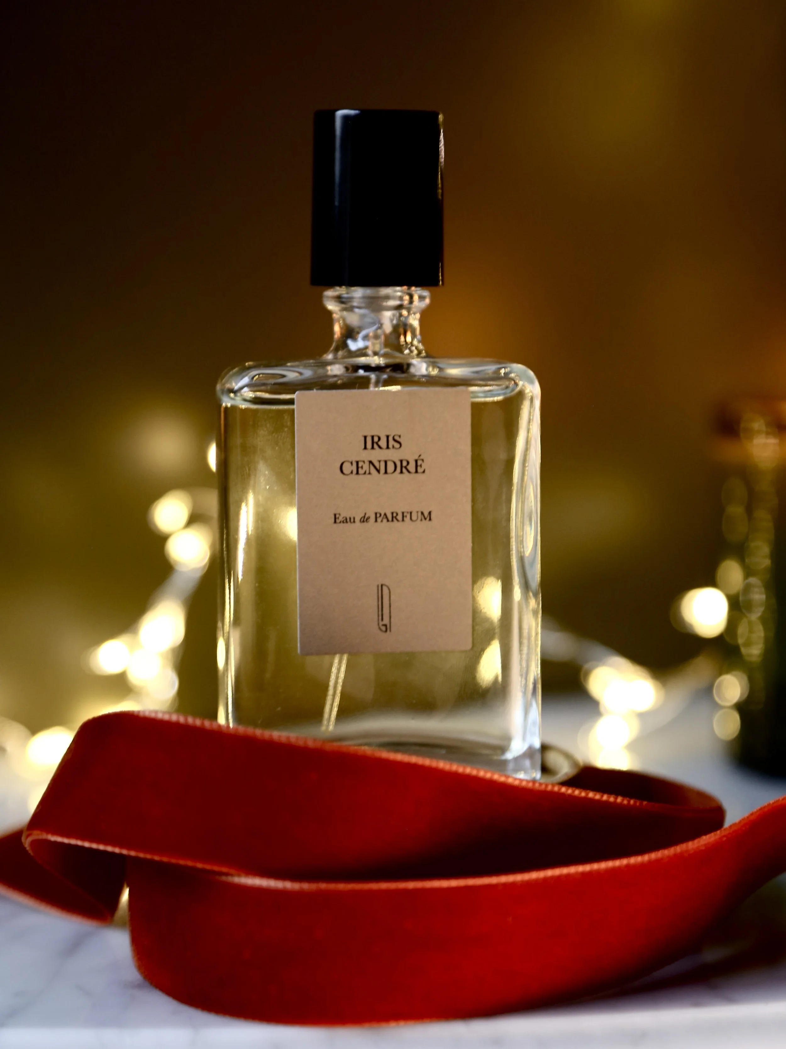 A clear glass perfume bottle with a black cap, labeled 'IRIS CENDRÉ Eau de PARFUM,' resting on a white surface with a red ribbon in front. Bokeh lights are blurred in the background.